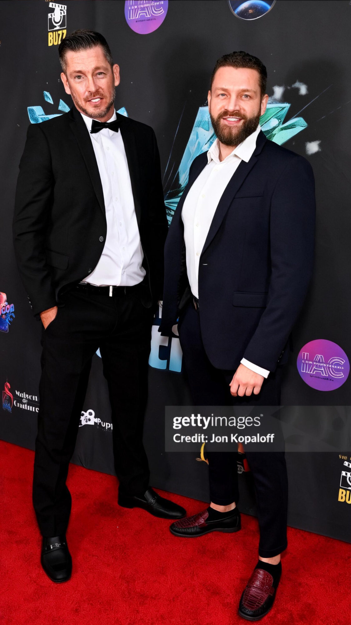 Two men in tuxedos standing on a red carpet at an event, smiling, with a dark background and event logos.