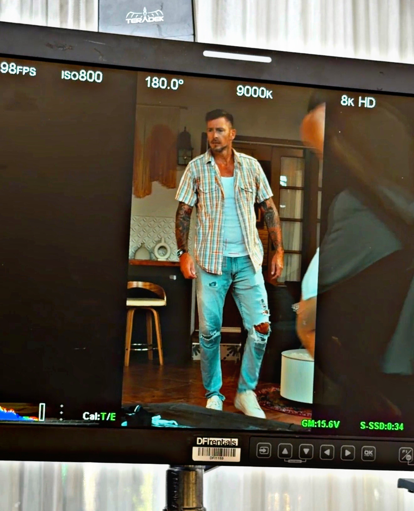 Man standing in a room with wooden furniture, wearing a plaid shirt and ripped jeans, reflected in a camera screen.