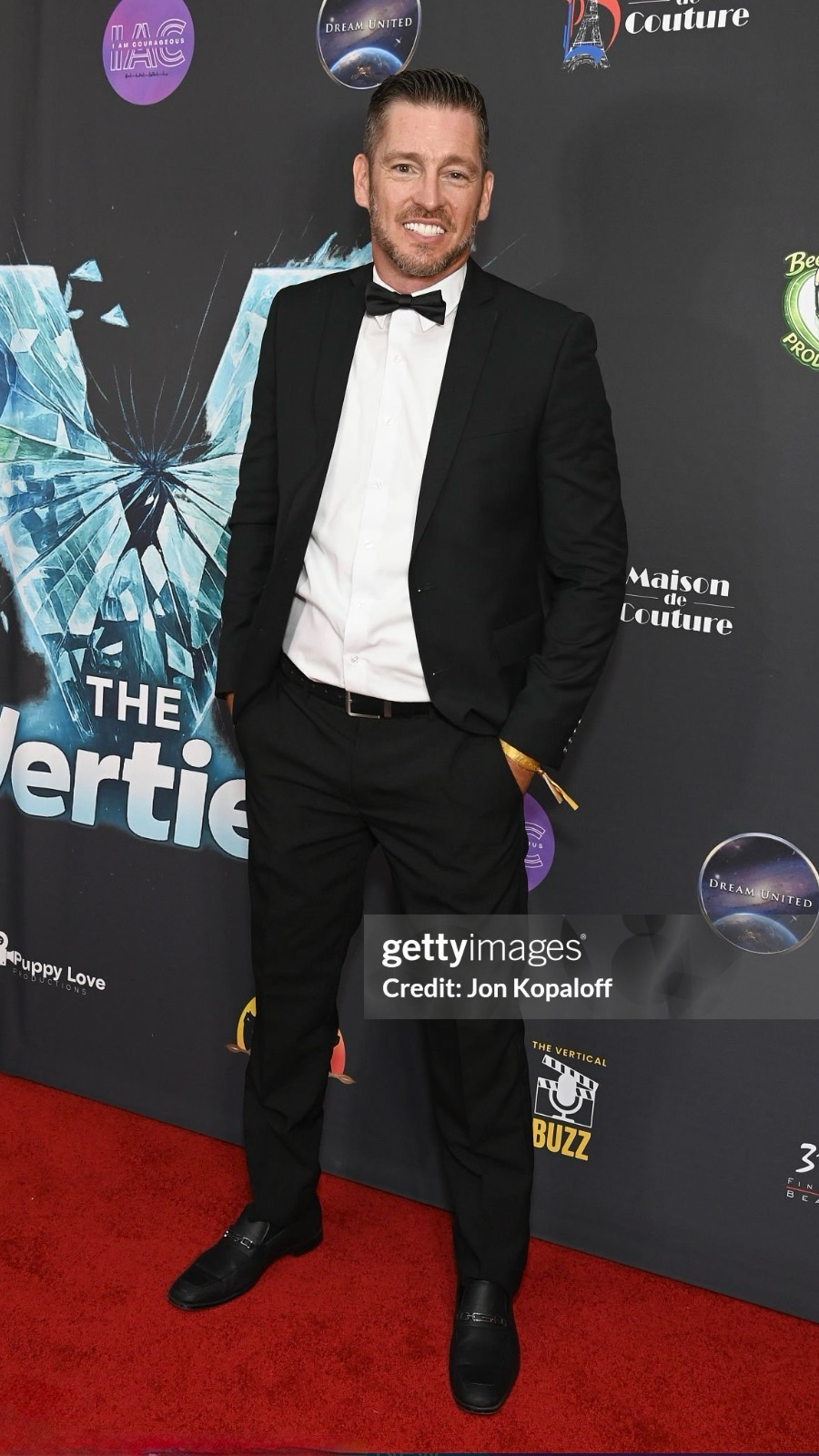 Man in tuxedo with bow tie standing on red carpet at event, smiling, background with logos and graphics.