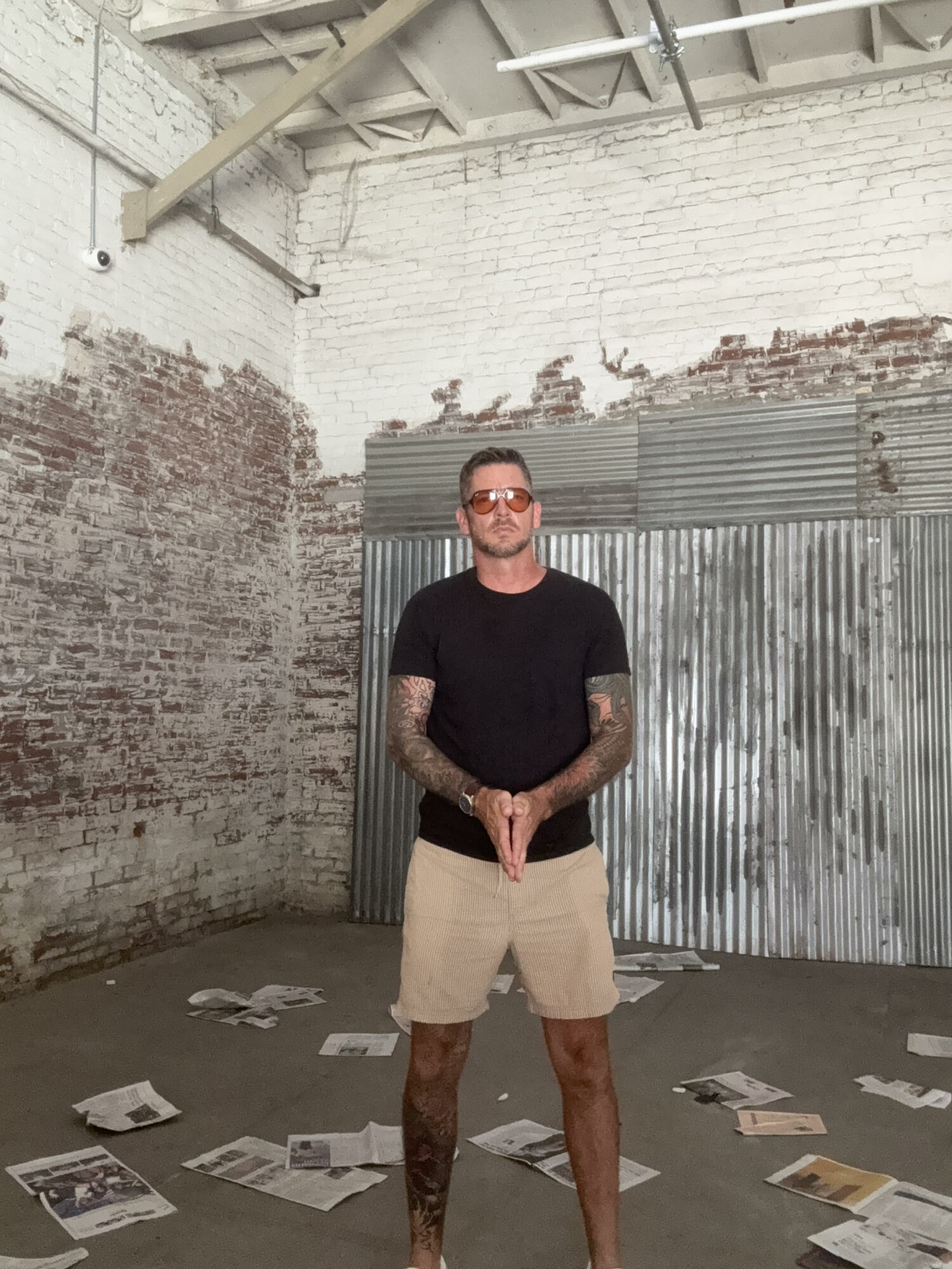 Man with tattoos wearing sunglasses, black t-shirt, and beige shorts standing in an industrial space with scattered papers.