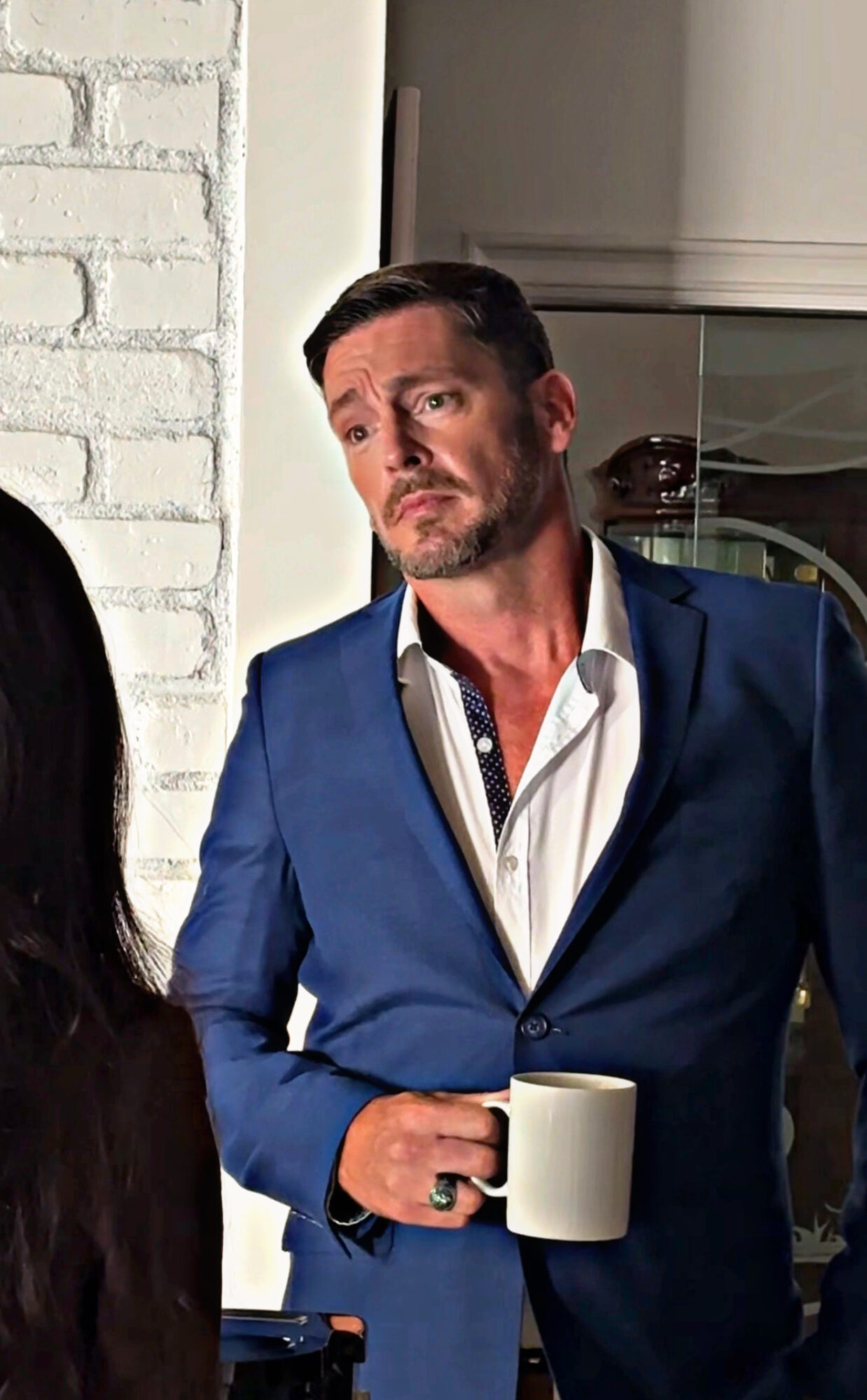 Man in a blue suit holding a white mug, standing indoors near a white brick wall.