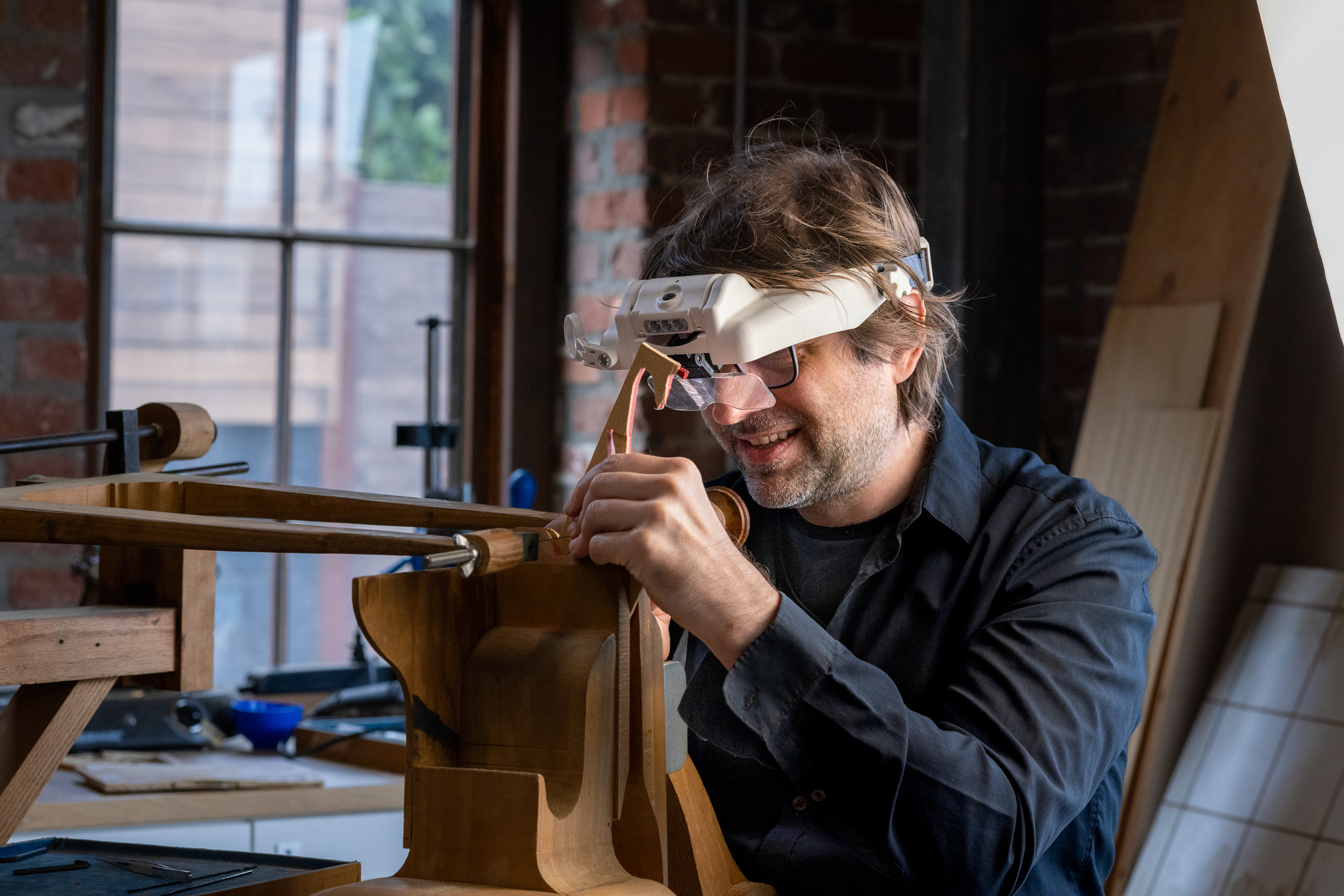 Man with glasses and beard working on a wooden guitar in a workshop, holding a tool near the instrument's body.