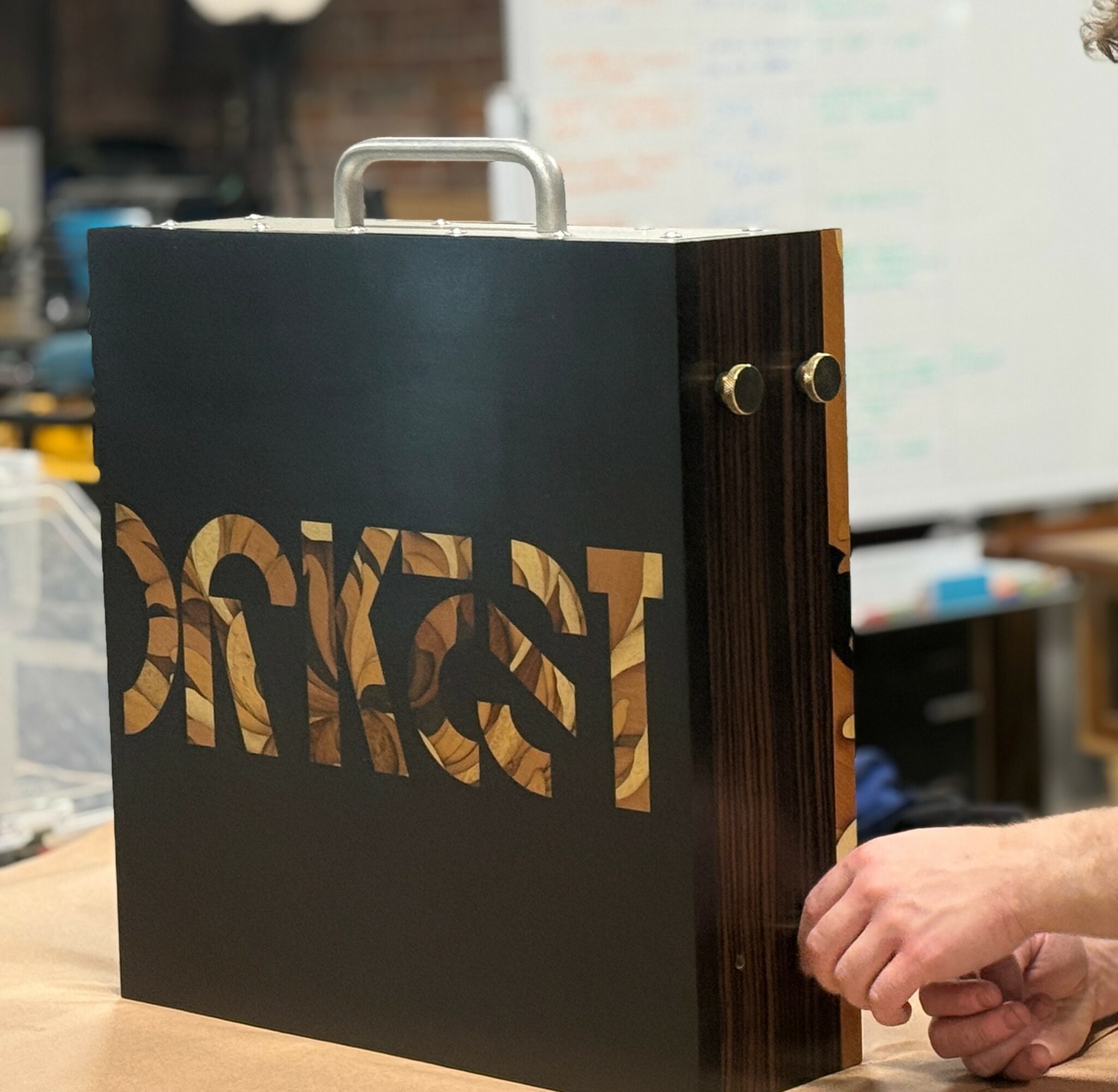 Person holding a black box with a wooden side, metal handle, and cut-out text design.