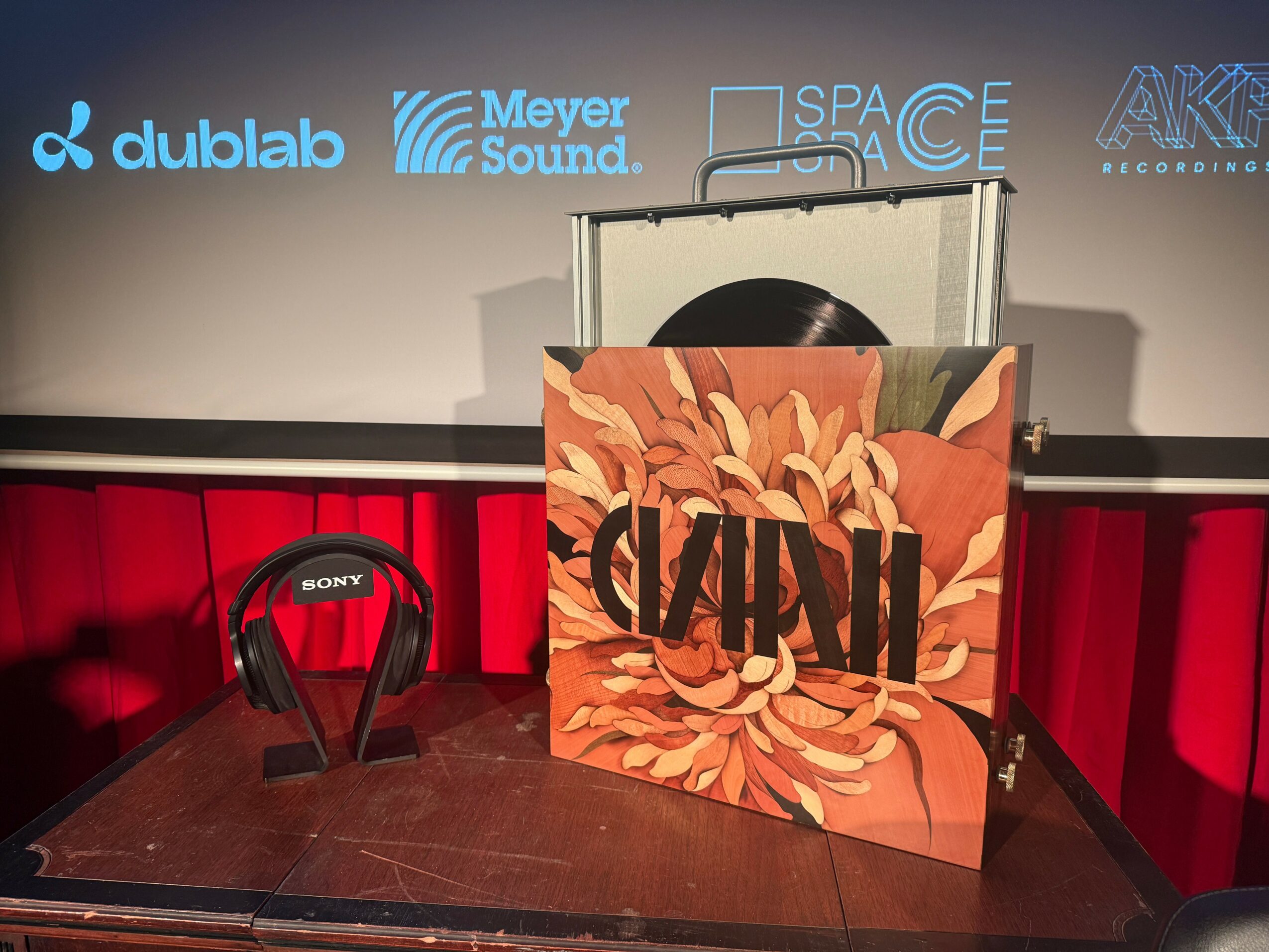 A large floral-patterned box with the word 'CASH' on it, placed on a wooden table with a Sony headphone set nearby, in a room with a screen displaying logos.