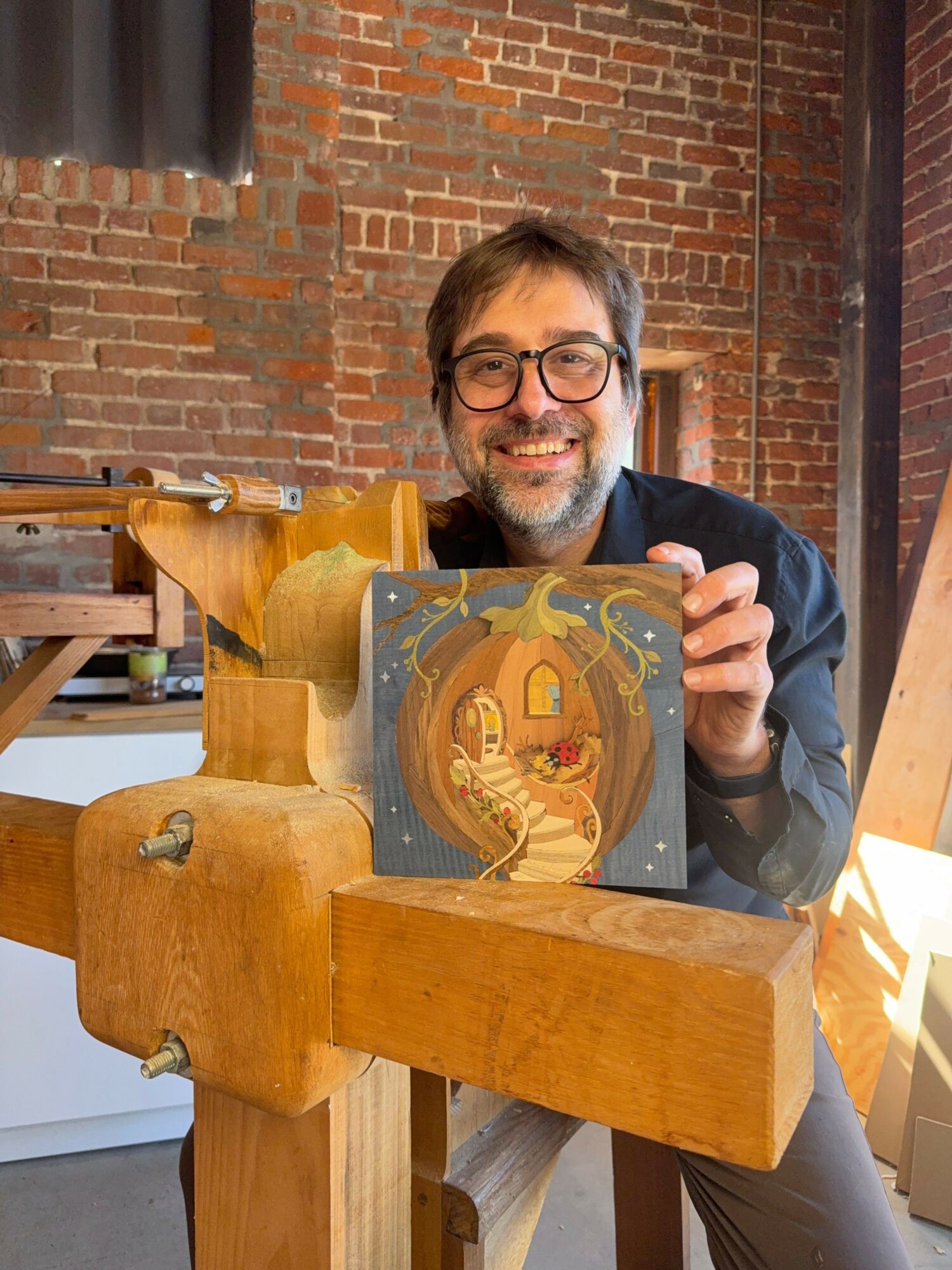 Man with glasses and beard looking at a painting on a wooden table, brick wall background.