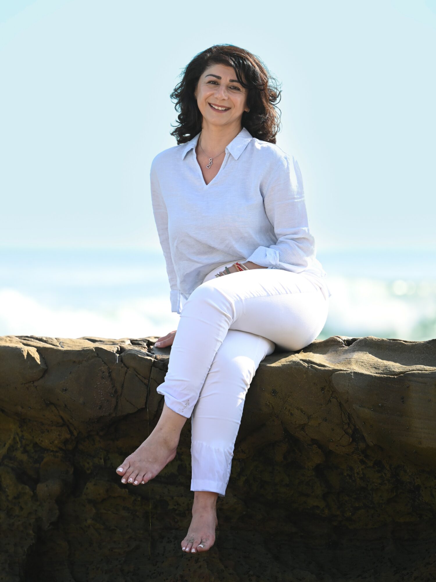 Woman sitting on a rock at the beach, smiling, with ocean and sky in background.