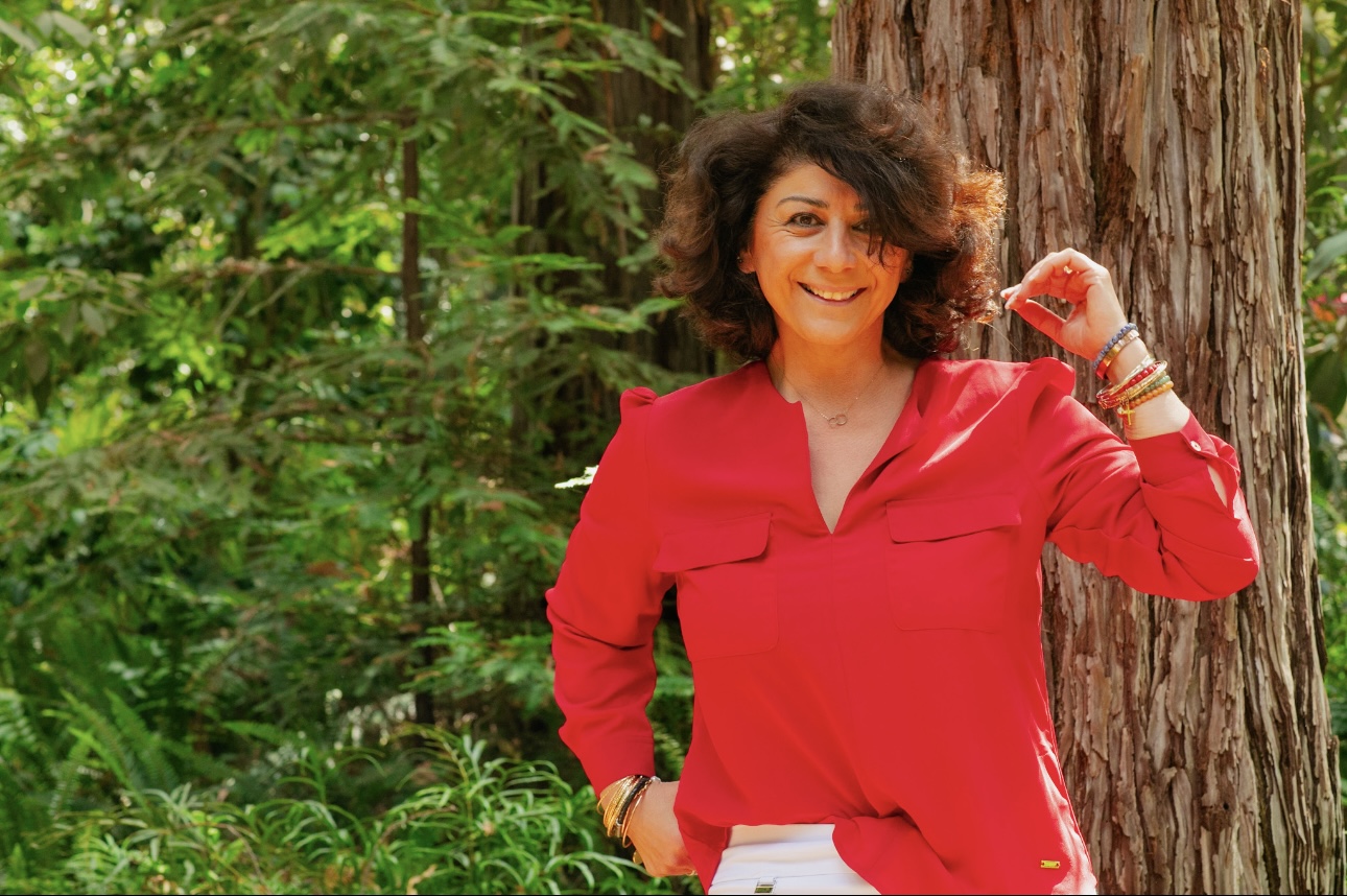 Woman with curly hair smiling outdoors, wearing a red blouse, standing next to a tree, surrounded by green foliage.