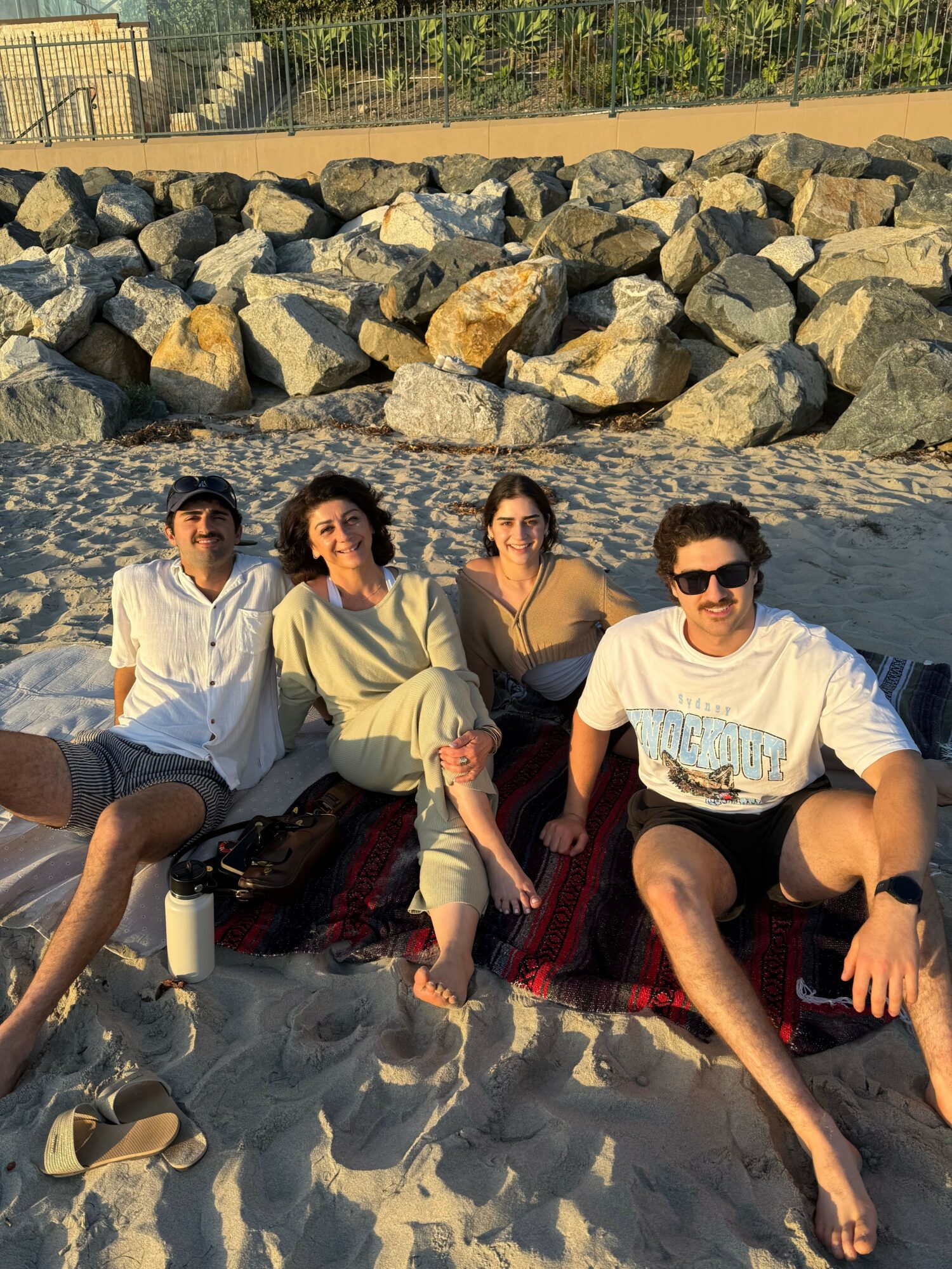Four people sitting on a sandy beach near rocks, with water and trees in the background, posing for a photo.