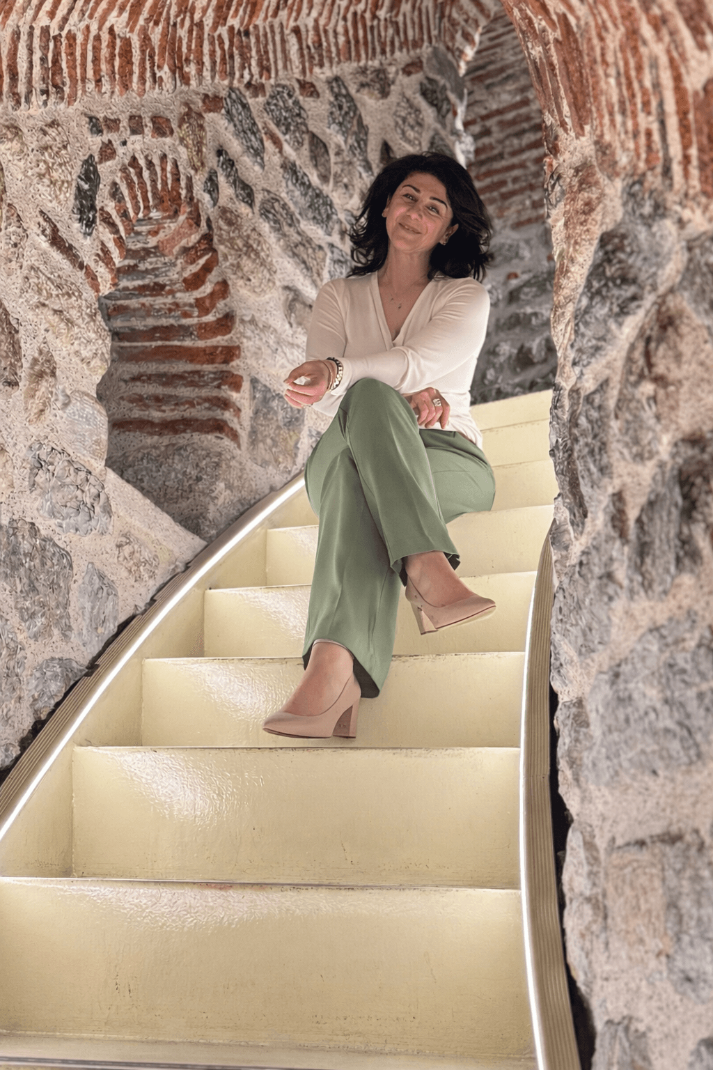 Woman sitting on illuminated staircase surrounded by stone walls, smiling, wearing a white top and green pants.