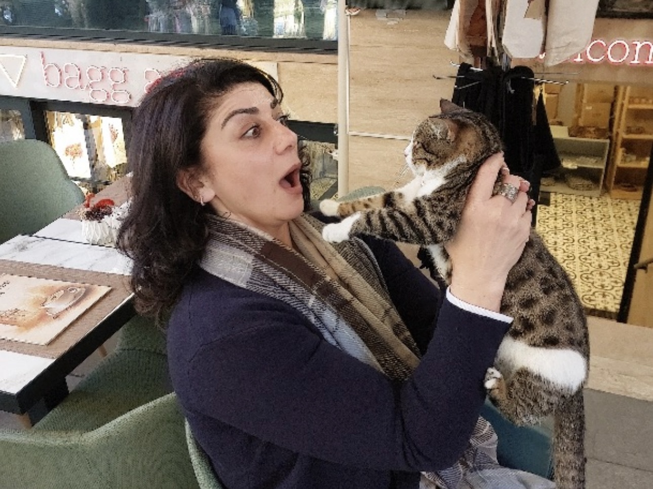 Woman holding a cat indoors, surprised expression, seated at a table with books and papers nearby.