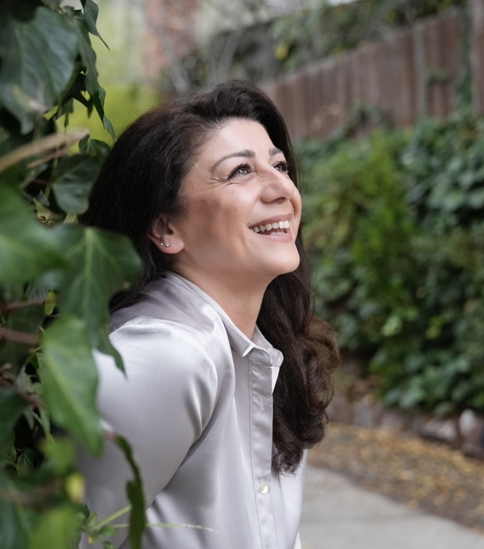 Woman with dark hair smiling outdoors, surrounded by green foliage and a garden path.
