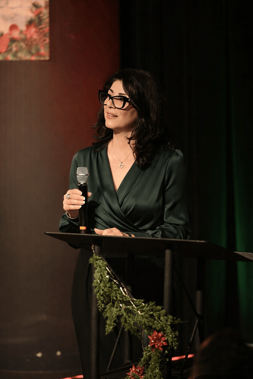 Woman with dark hair and glasses speaking into a microphone on stage, wearing a dark green satin blouse, with a black curtain background.