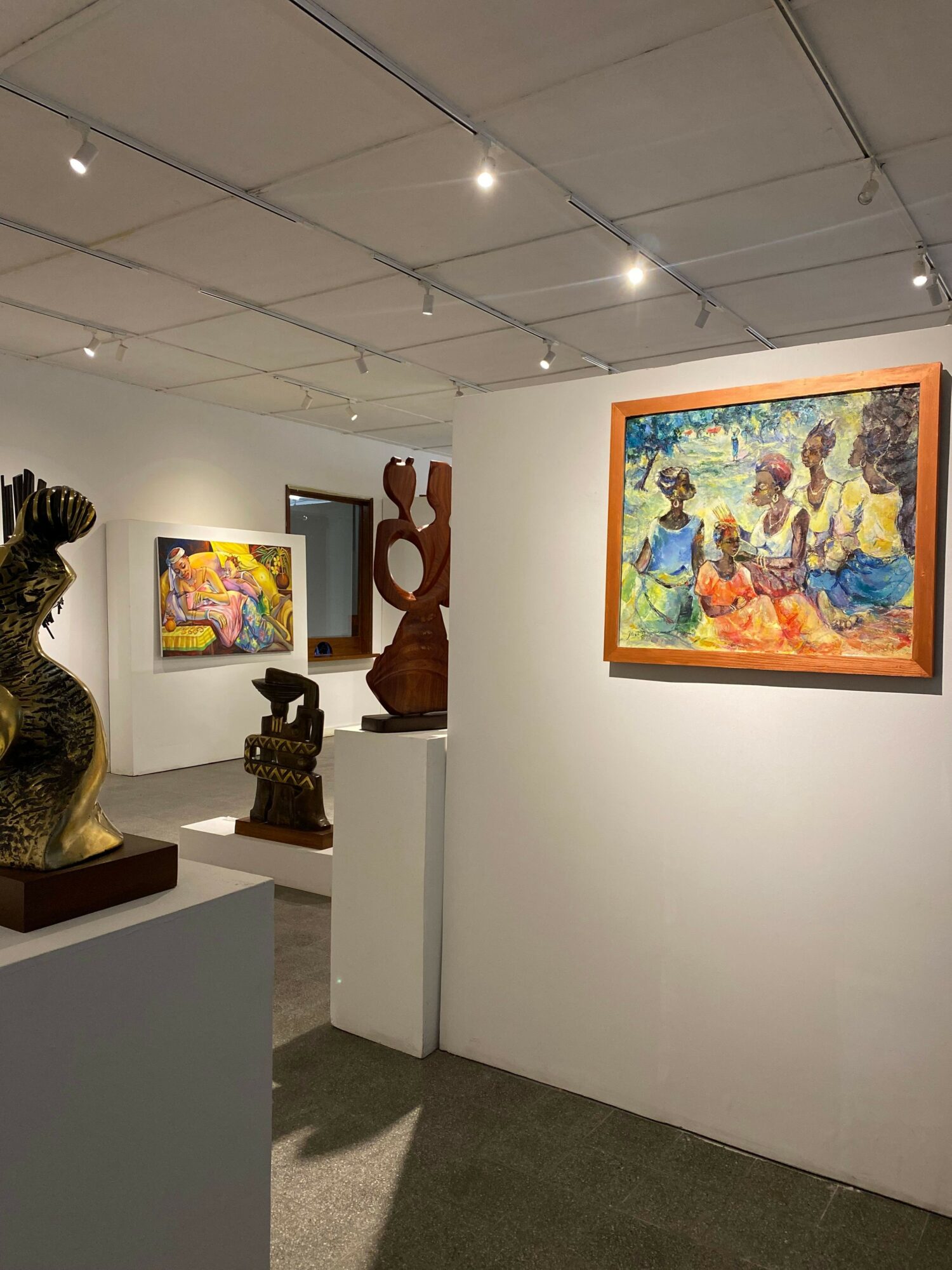 Gallery with colorful paintings on white walls, sculptures on pedestals, and a tiled ceiling with lights.