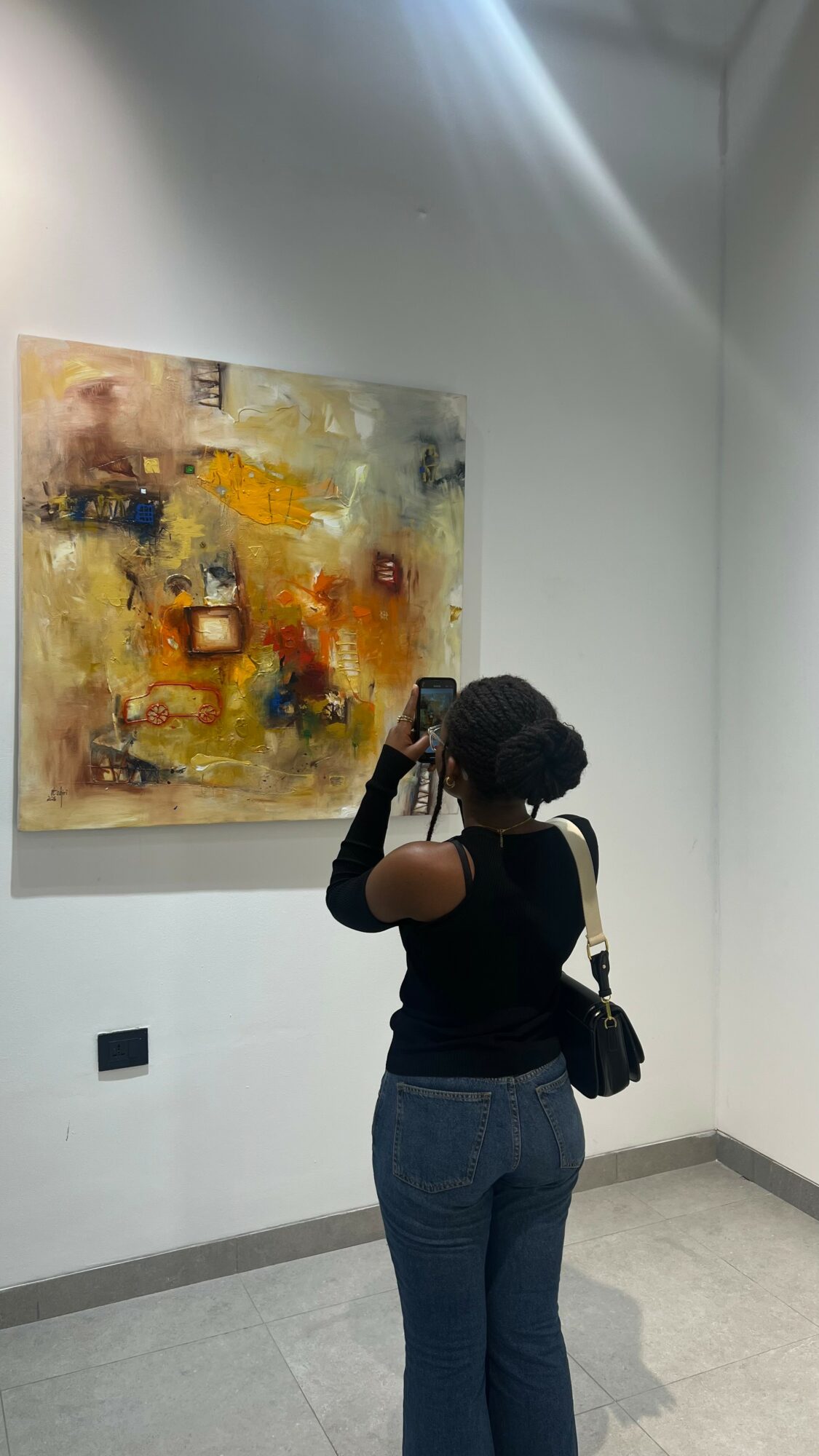 Person taking photo of abstract painting in gallery, standing with back to camera, wearing black top and jeans, carrying a bag.