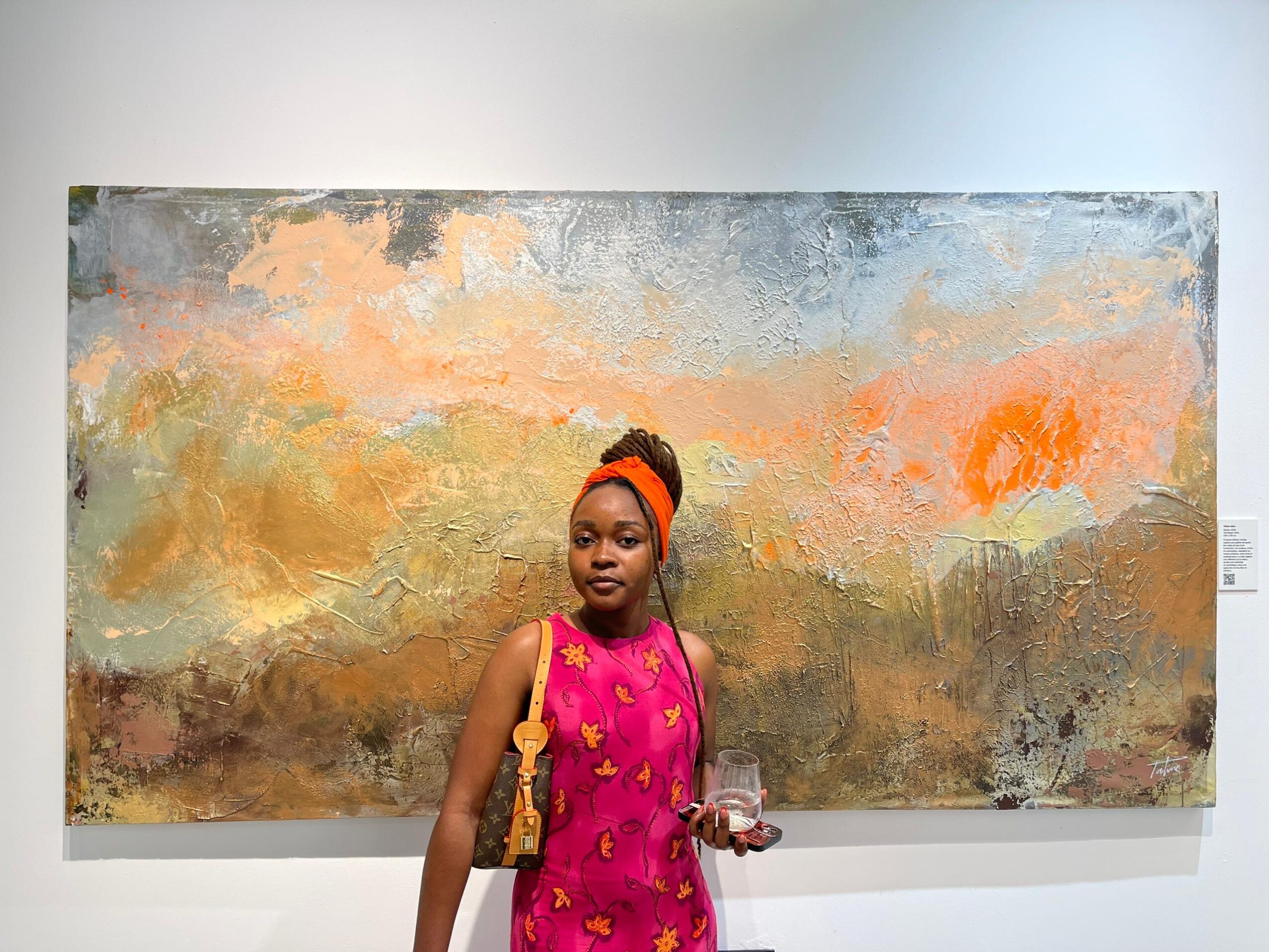 Young girl in pink dress with orange headband holding a drink, standing in front of an abstract landscape painting.