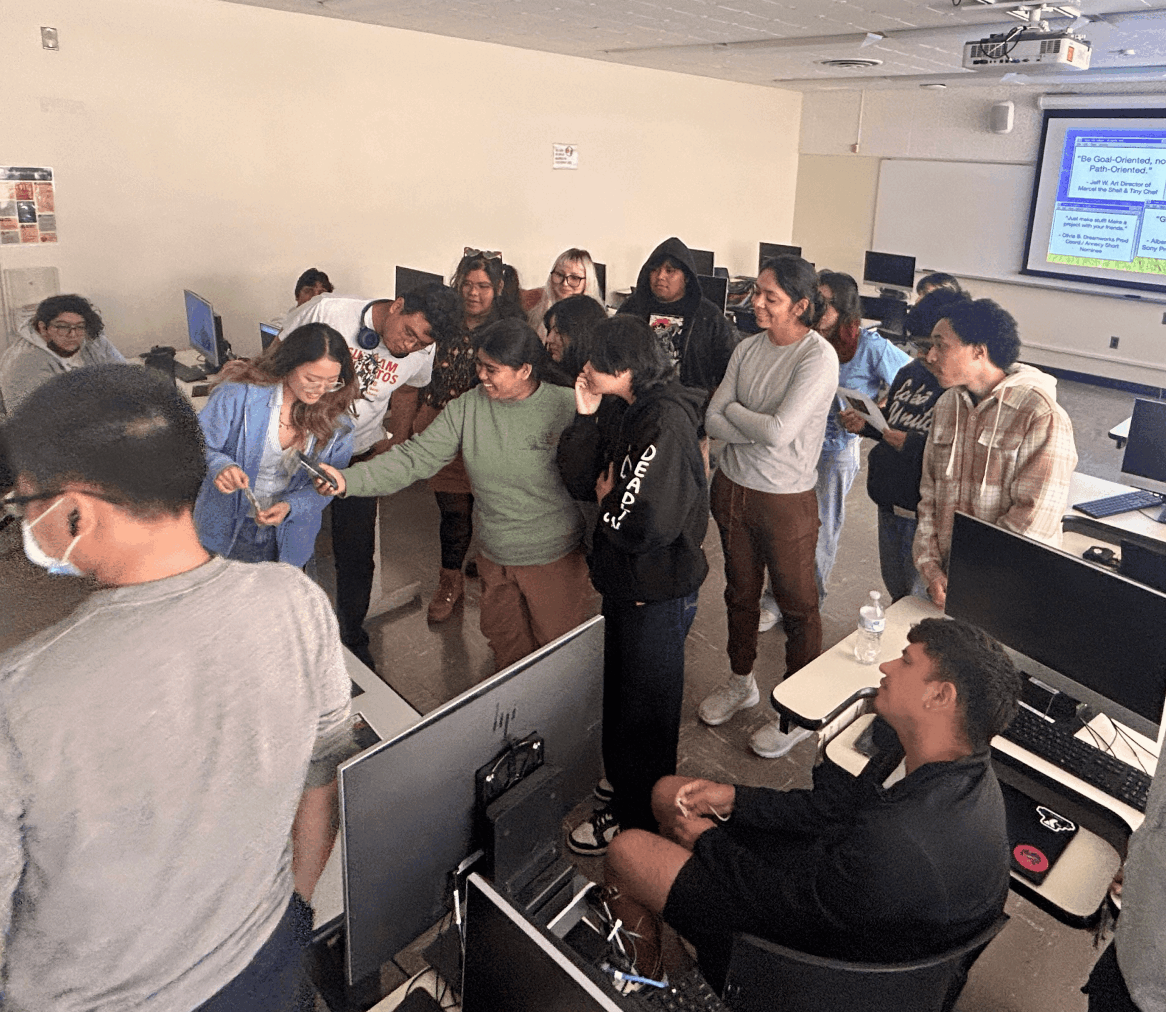 Group of students in a classroom, some standing and some seated, with a projector screen displaying a presentation.