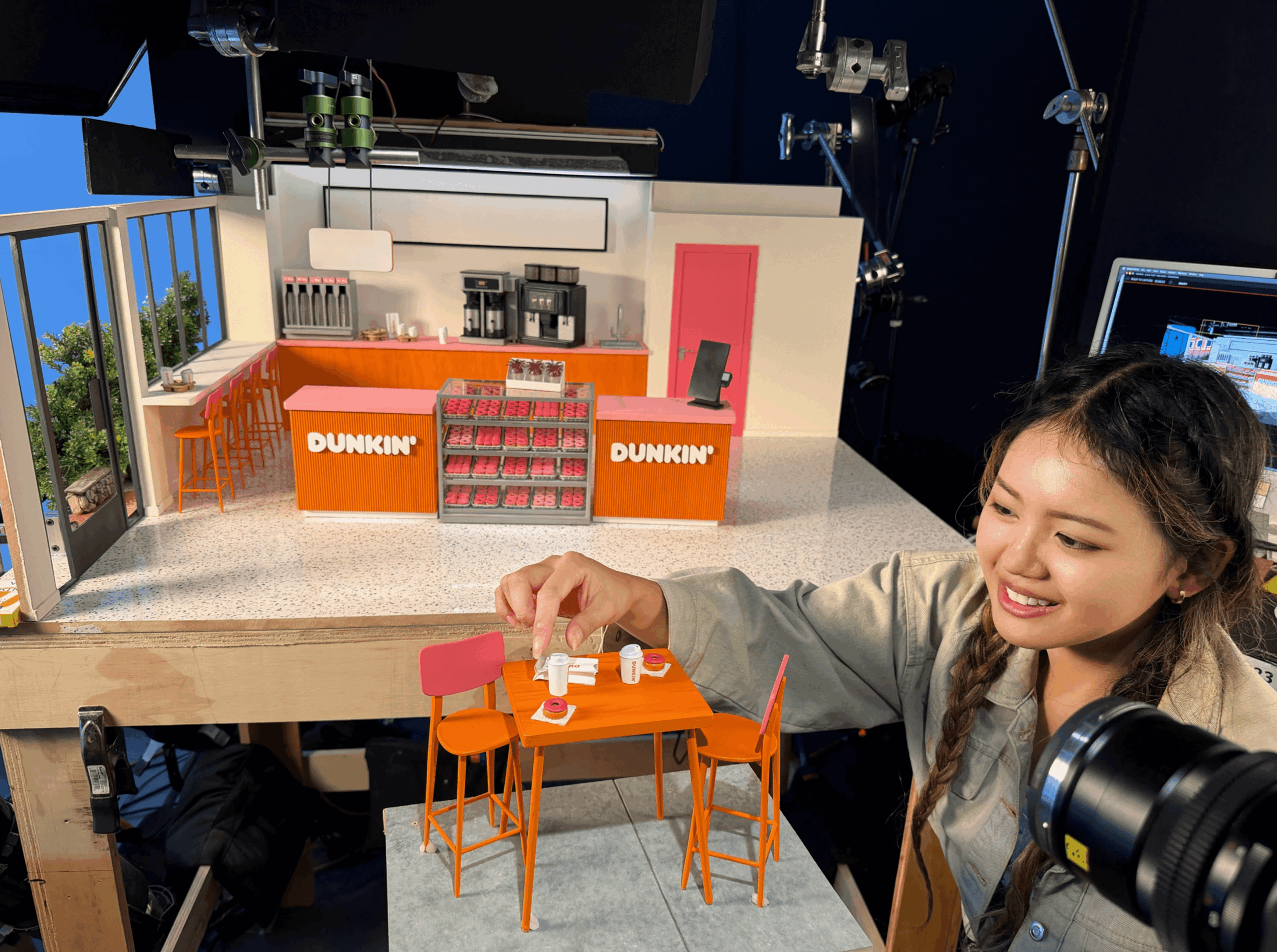 Young woman smiling at a miniature orange table with chairs, reaching for a small object, in a studio setting.