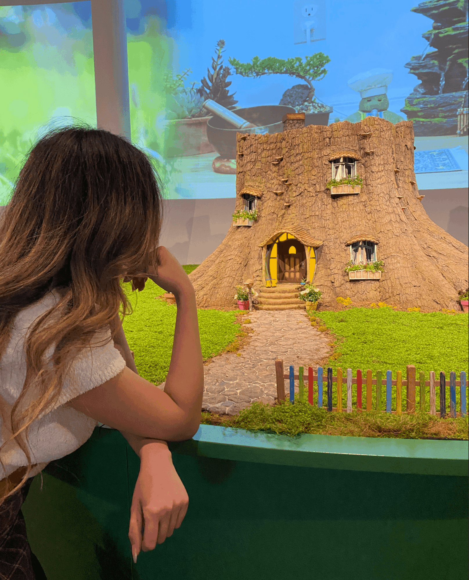 Girl with long hair looking at a model of a castle with a colorful fence, displayed indoors with a nature scene background.
