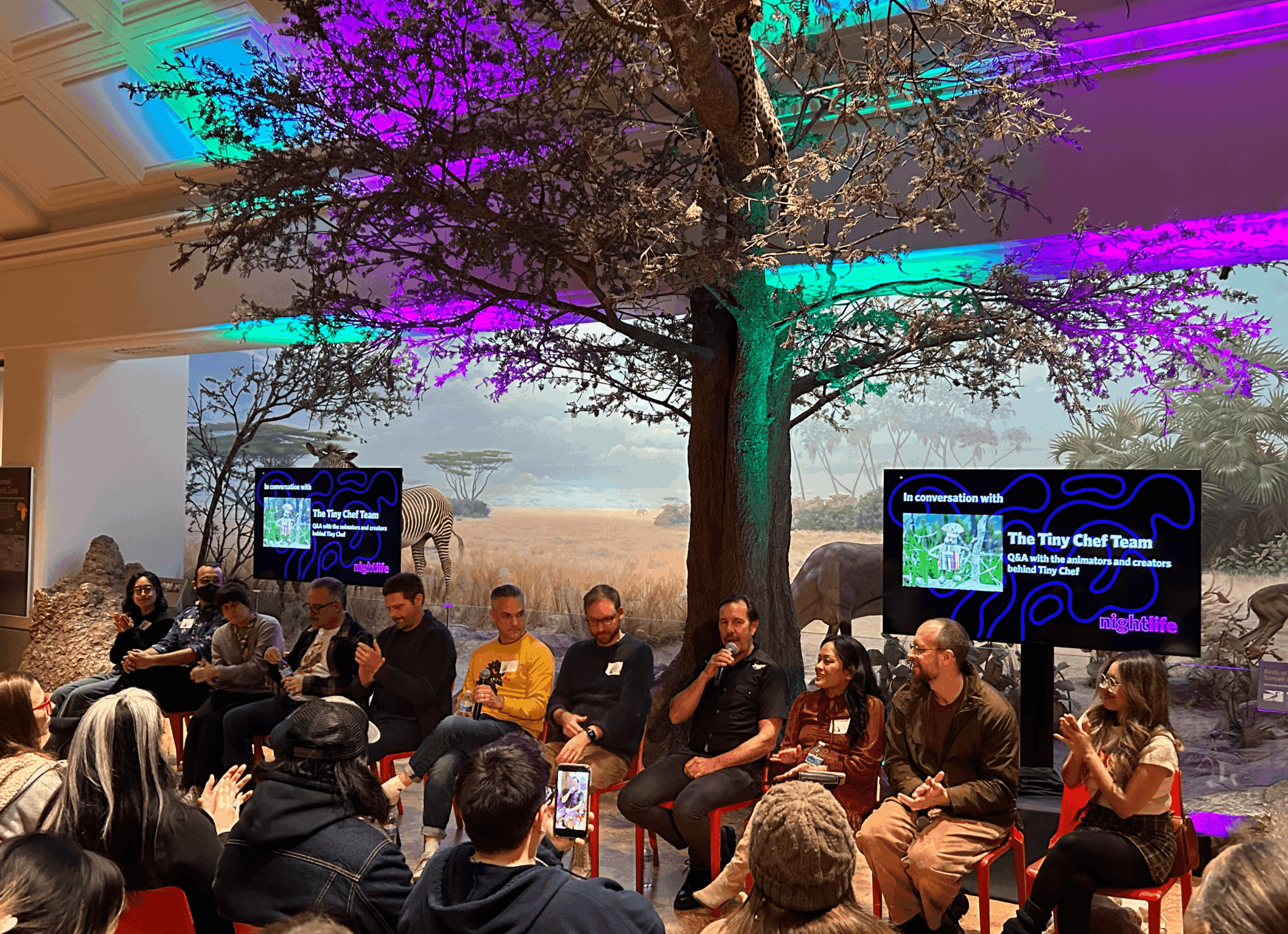 Panel discussion with people seated in front of a large tree display and screens showing event information.