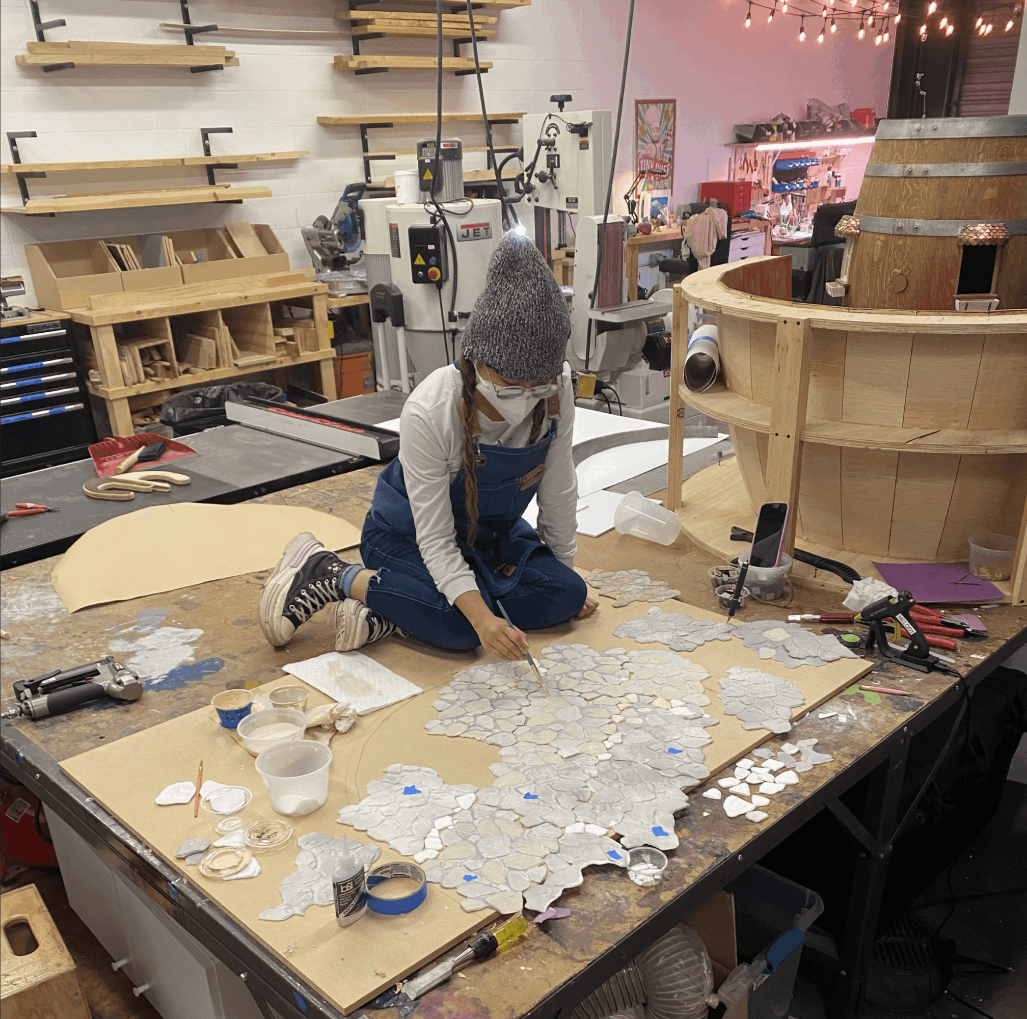 Person working on a large wooden project in a workshop, surrounded by tools and materials, sitting on the floor.