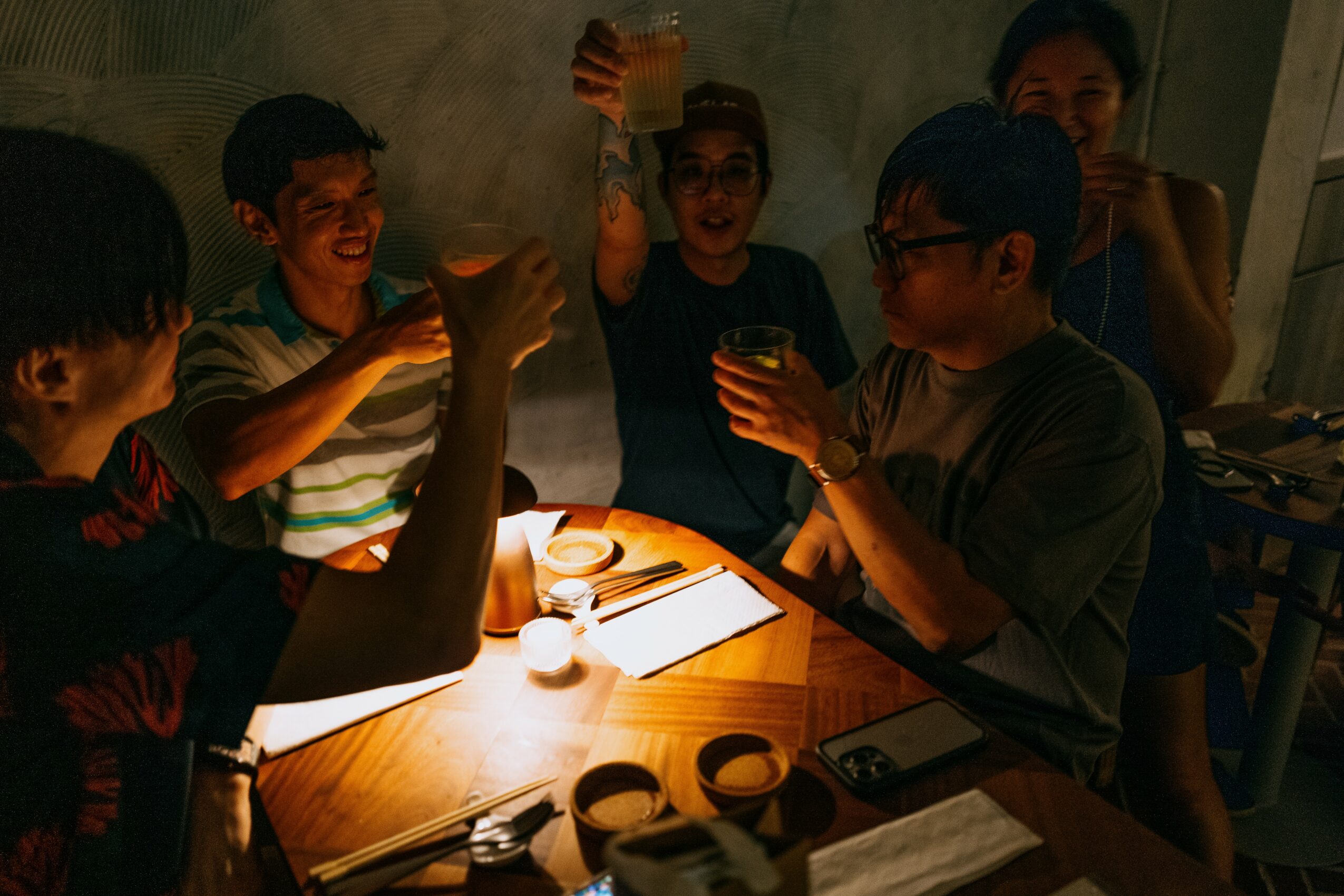 Group of five people sitting around a table, raising glasses in a toast, with a lit candle and notebooks on the table.