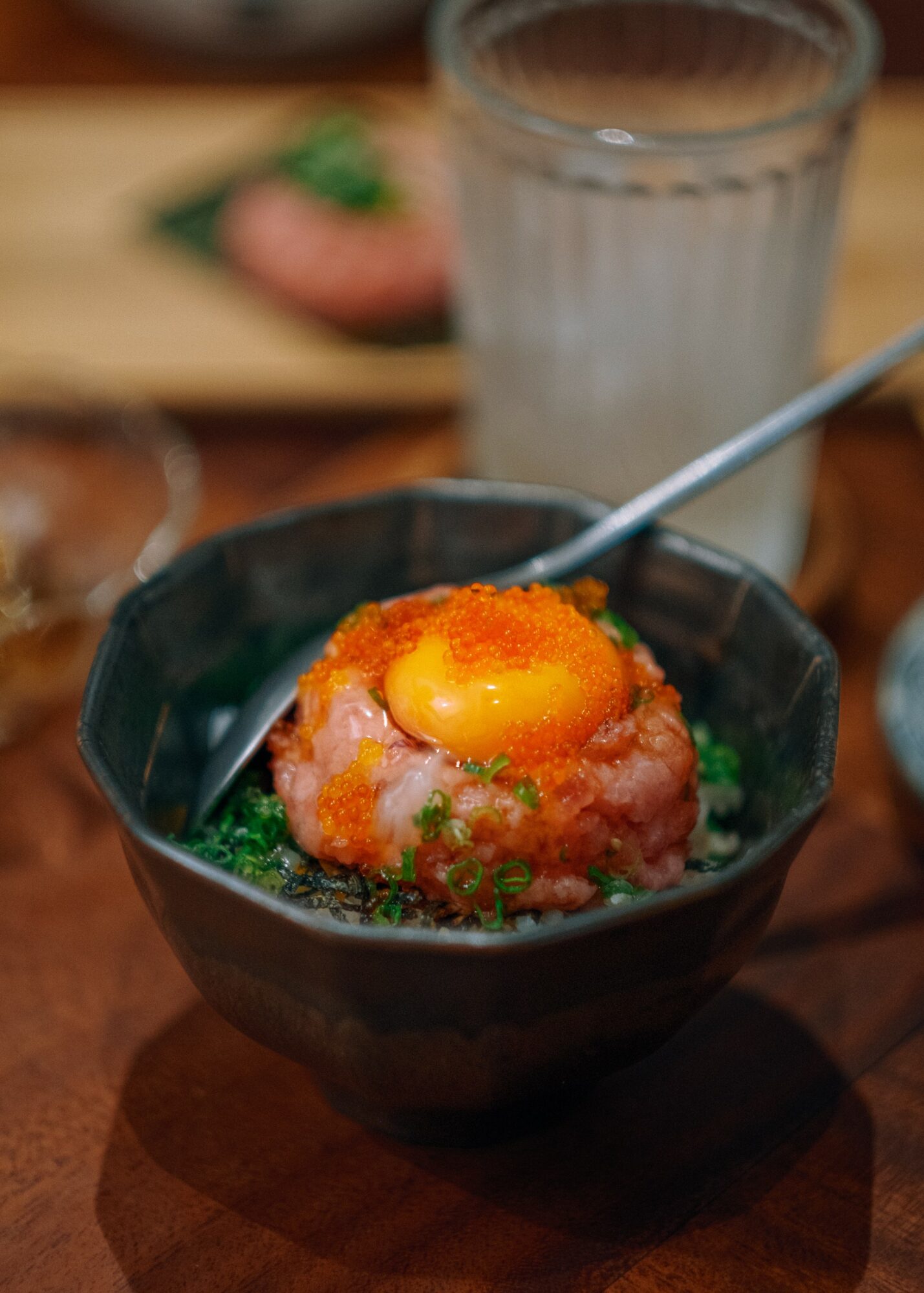 A bowl with raw minced meat topped with an egg yolk and green herbs, with a spoon inside, on a wooden table.