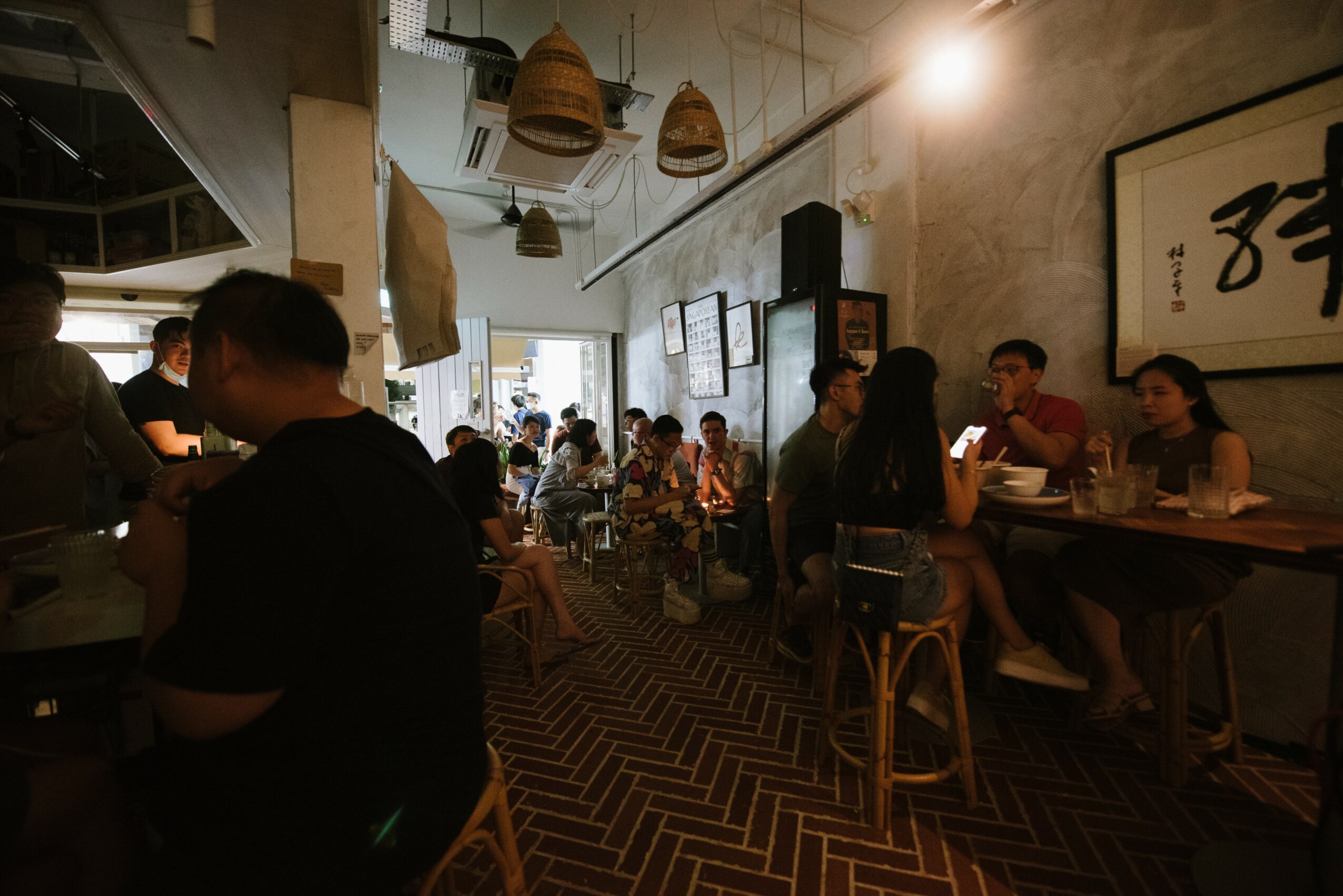 People sit at tables and a bar in a cozy, dimly lit cafe with hanging lights and artwork on the wall.