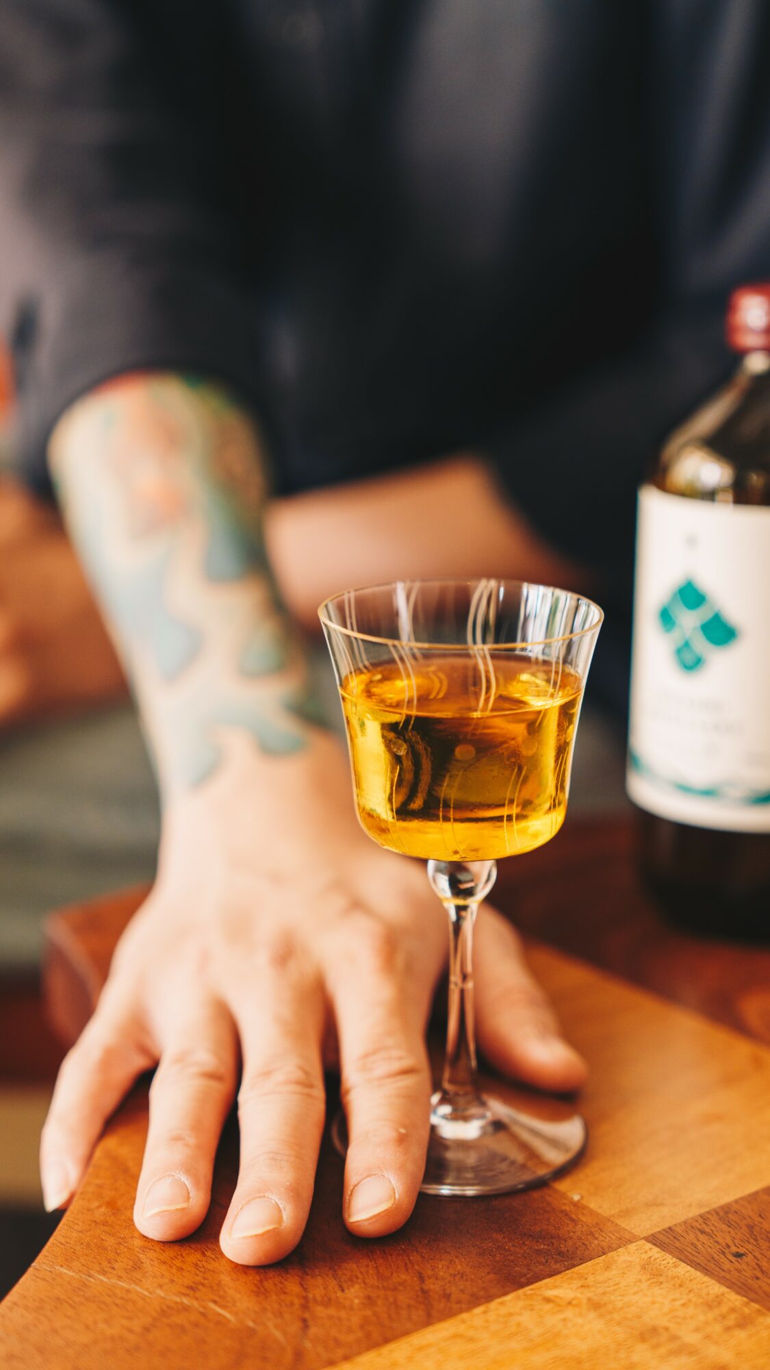 Person's hand resting on a wooden table with a glass of amber-colored drink, blurred background with a bottle, and tattooed arm