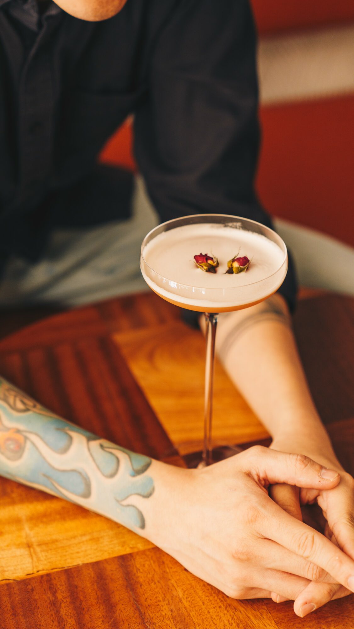 Person with tattooed arm sitting at a wooden table, holding hands with another person, with a cocktail glass on the table.