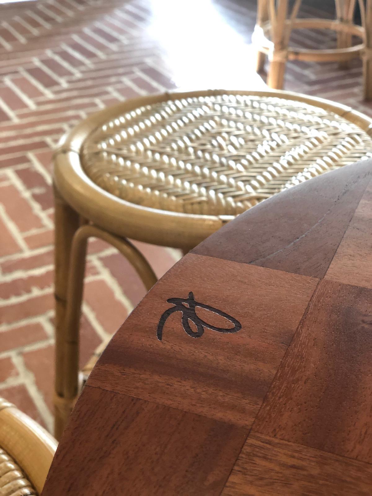 Close-up of a wooden table with a black signature or mark, and a woven rattan chair in the background, brick floor visible.