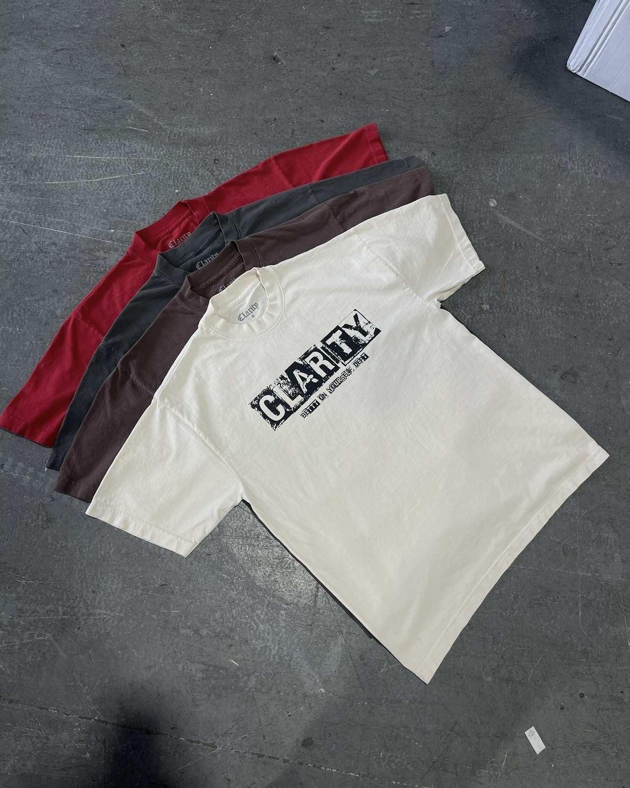 Four folded T-shirts in red, brown, black, and white with text, on a gray surface.