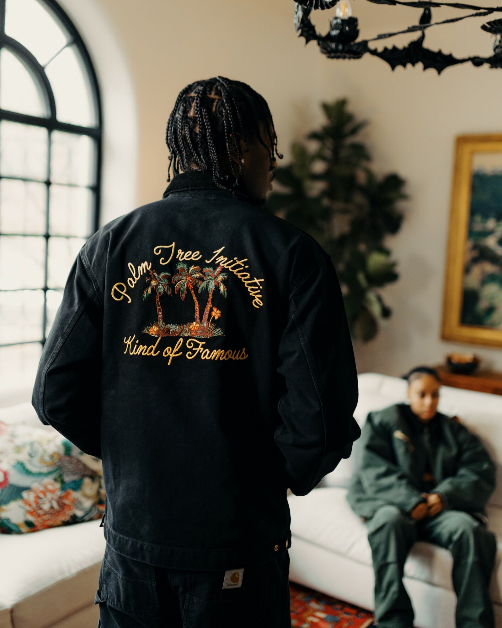 Person with braided hair wearing a black jacket with embroidered text and a palm tree design, standing indoors near a window.