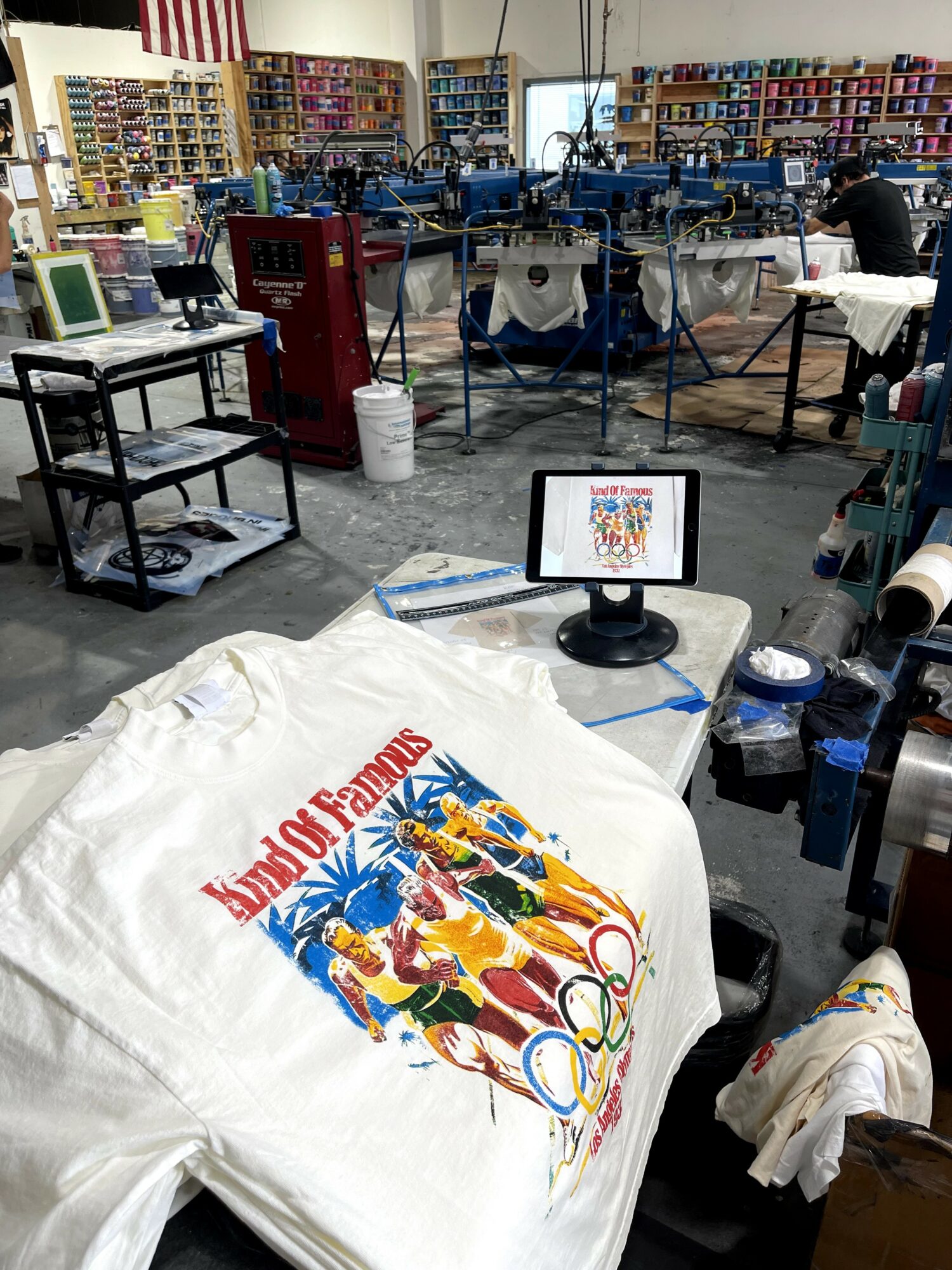 Indoor workspace with printing equipment, a computer monitor displaying a colorful design, and a fabric with a vibrant print, surrounded by supplies.