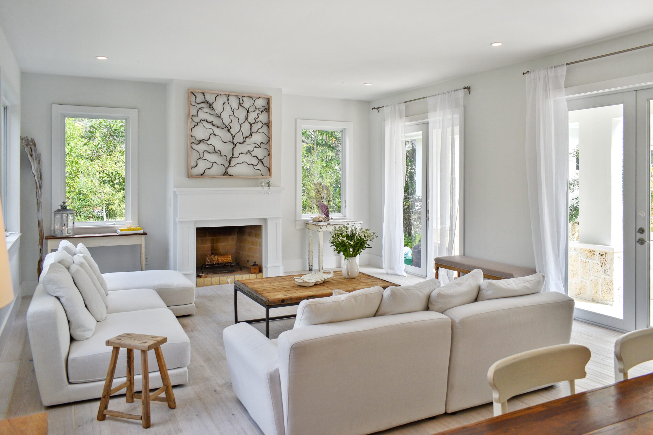 Bright living room with white sofas, a wooden coffee table, fireplace, and large windows with curtains, overlooking greenery.