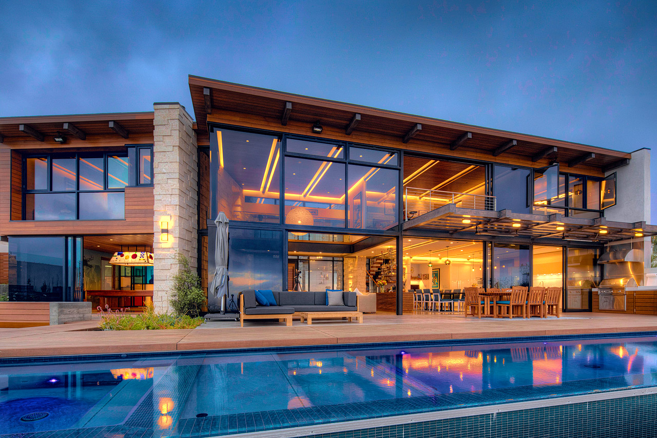 Modern building with large glass windows, outdoor seating, and a swimming pool in front, under a cloudy sky.