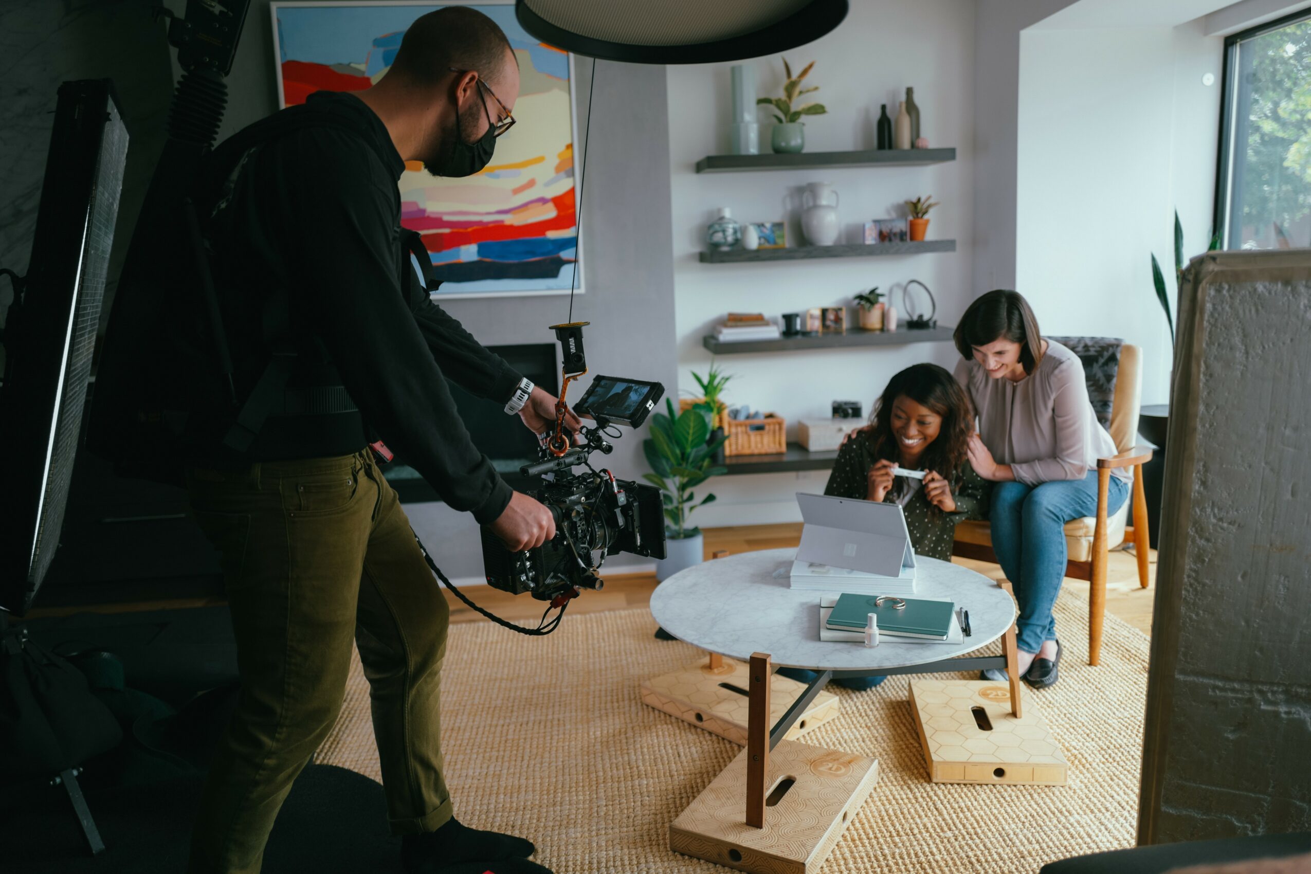 Two women sit on a couch using a laptop, while a man films them with a camera in a bright living room.