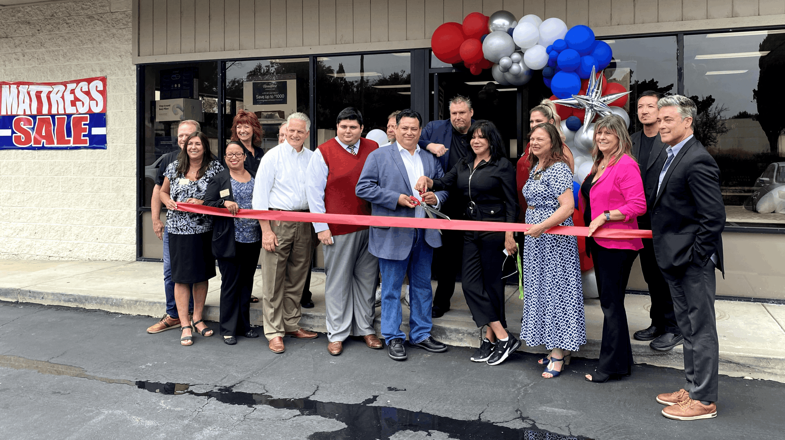 Group of people at a ribbon-cutting event outside a store with balloons and a sale sign, celebrating a grand opening.