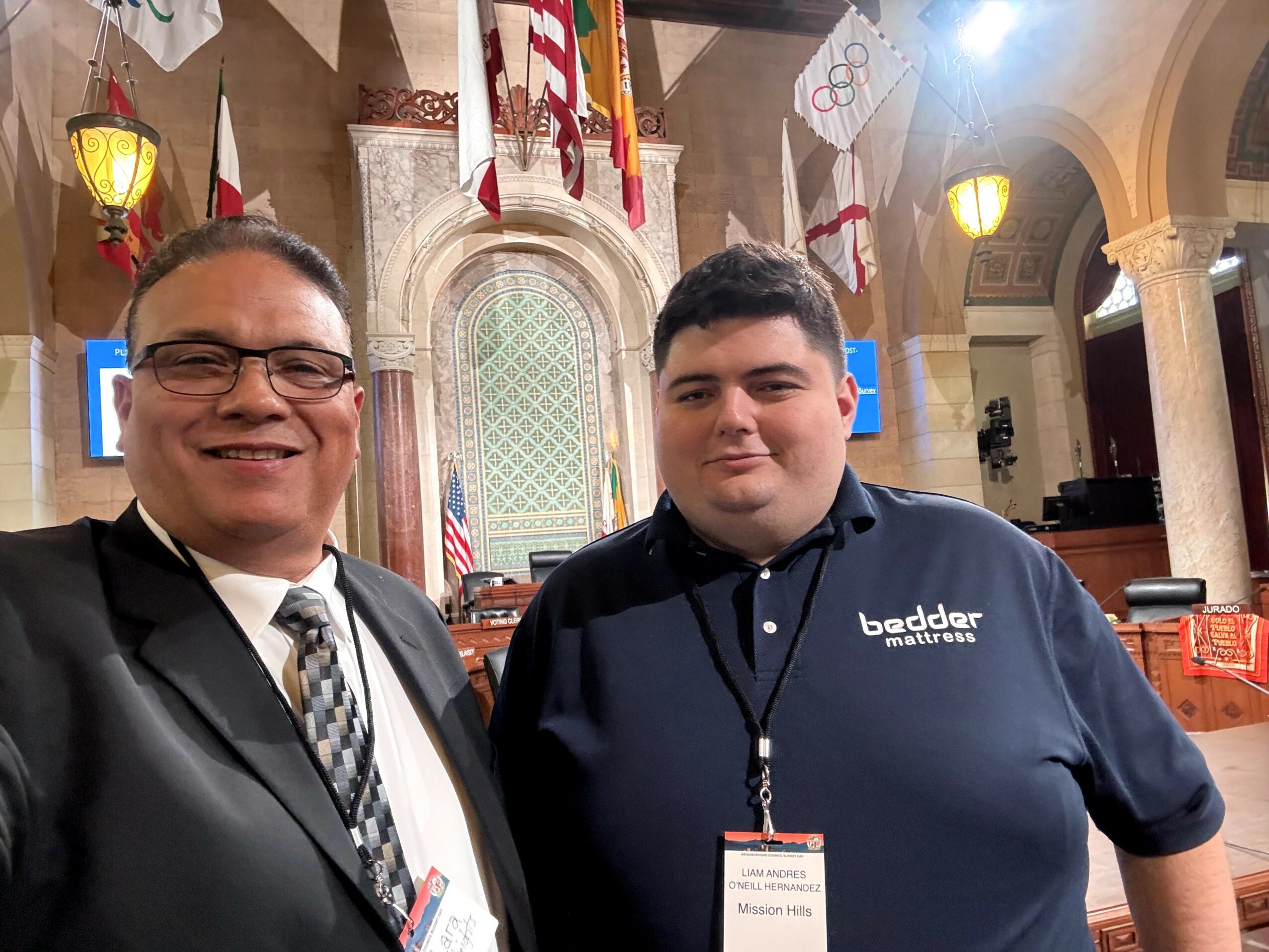 Two men standing inside a decorated hall with flags and stained glass window behind them.