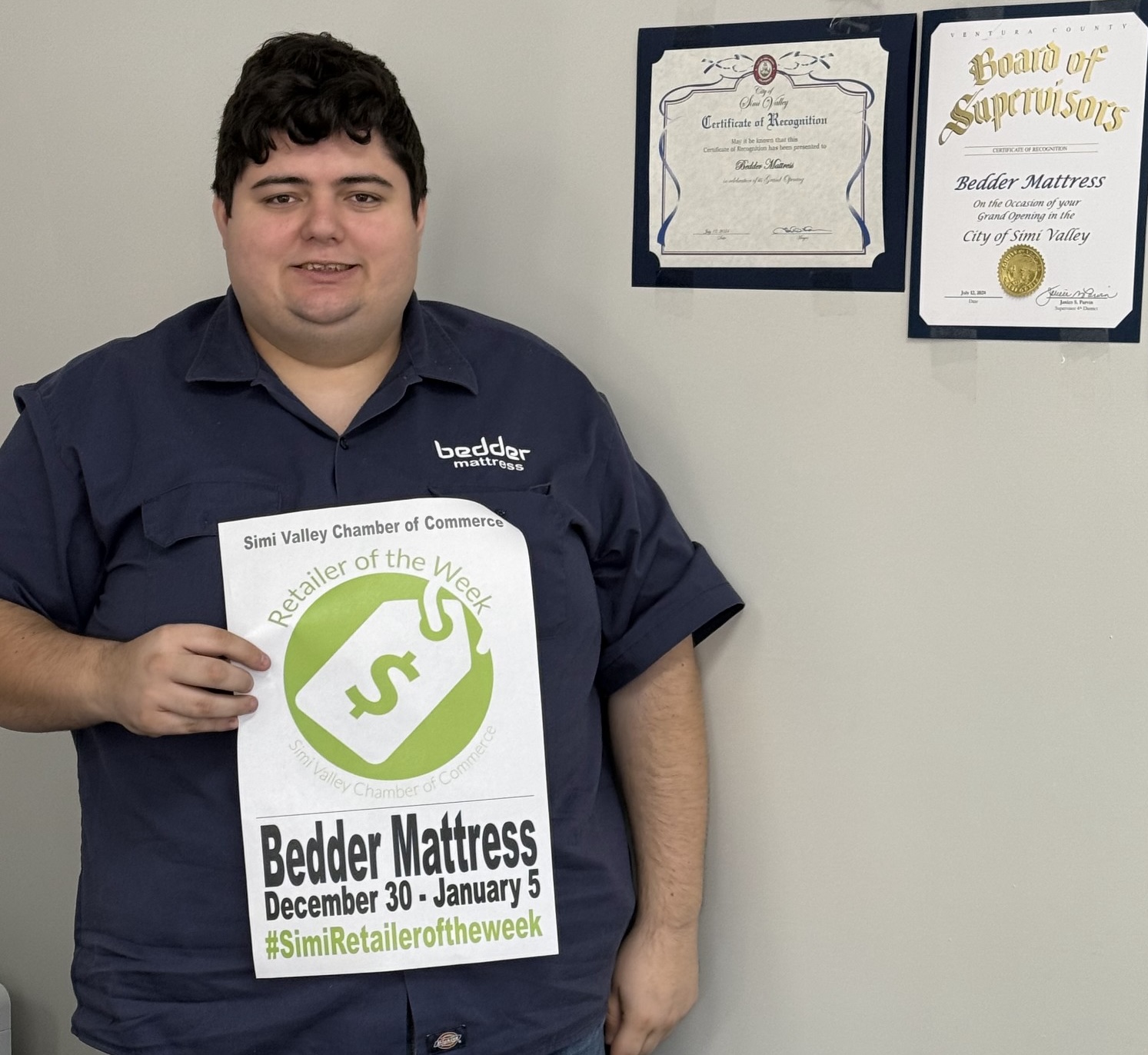 Young man holding a Bedder Mattress sign, standing in front of certificates on a wall.
