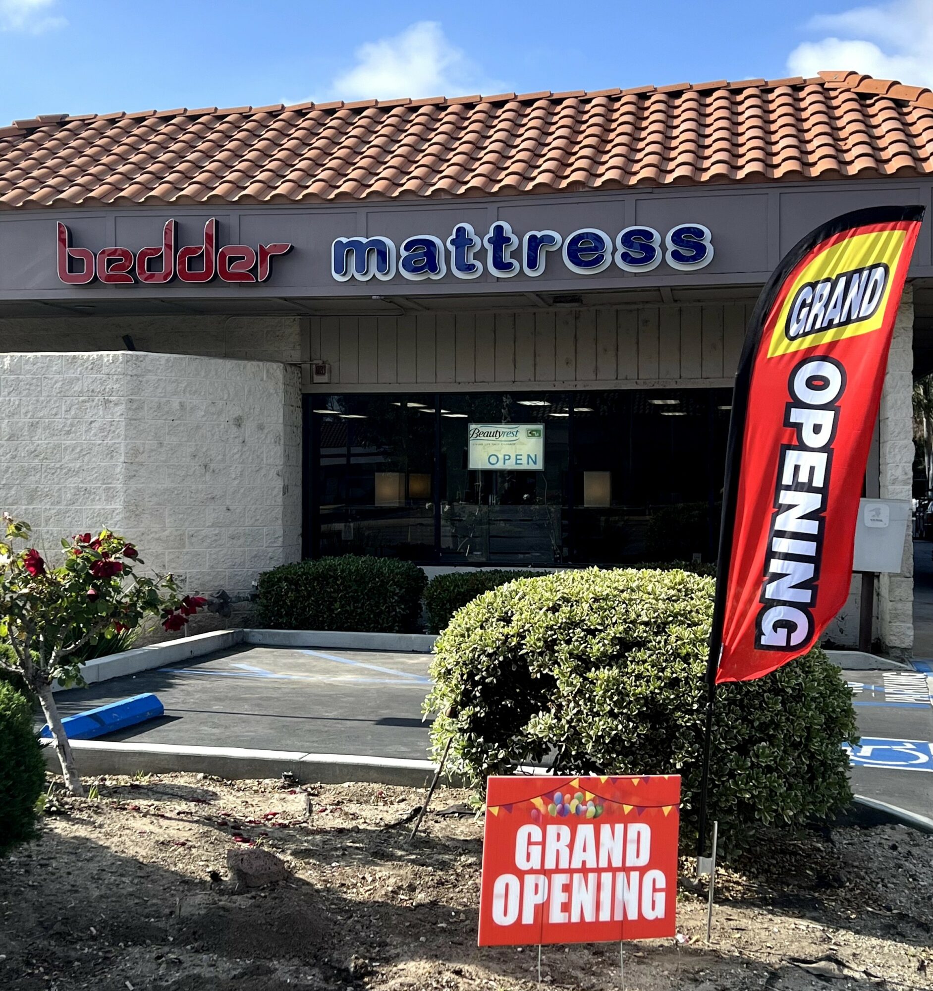 Store with a sign indicating grand opening, bushes, and a parking lot in front.