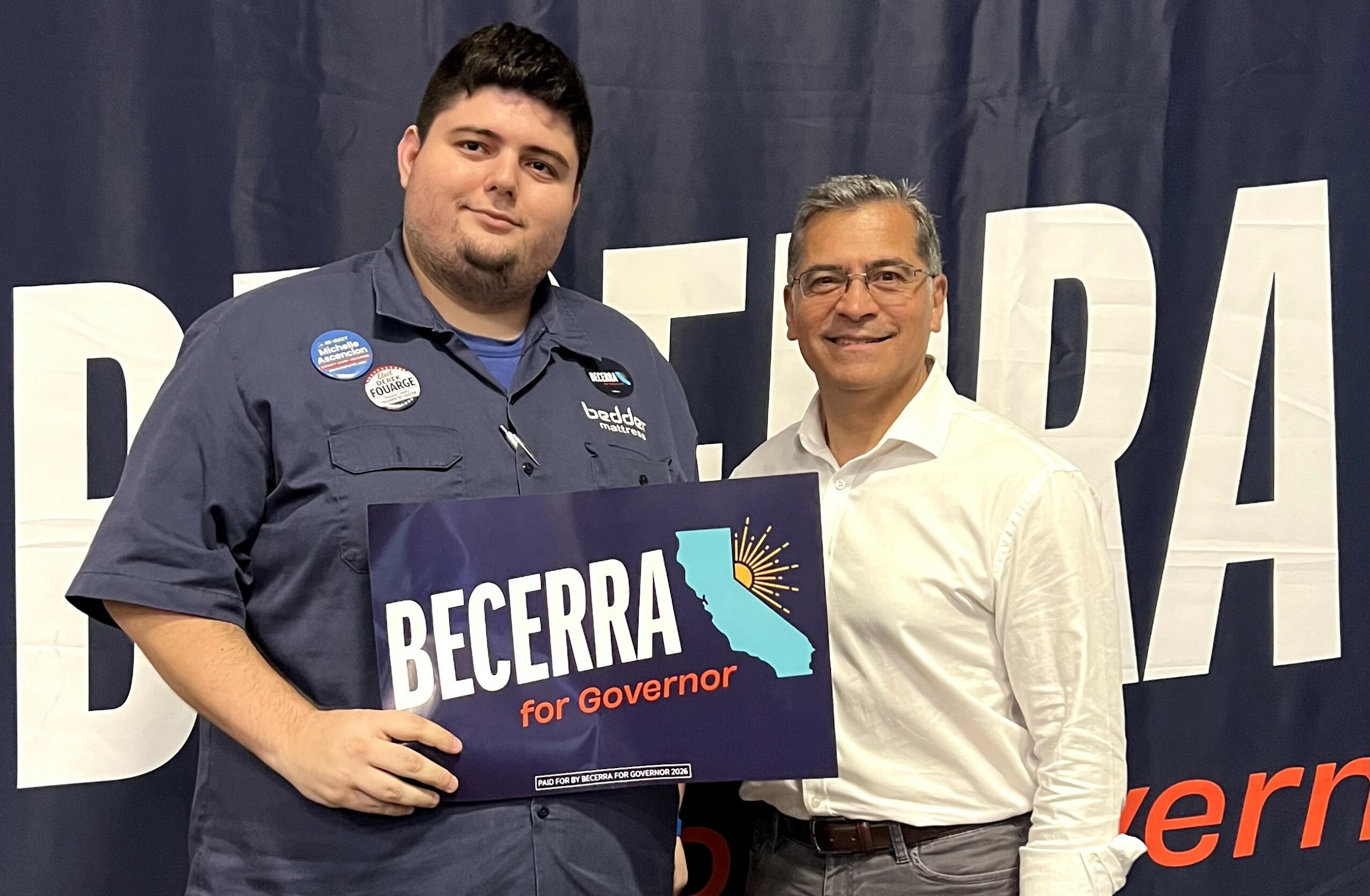 Two men standing together, one holding a sign that says 'BECERRA for Governor,' in front of a dark curtain backdrop.