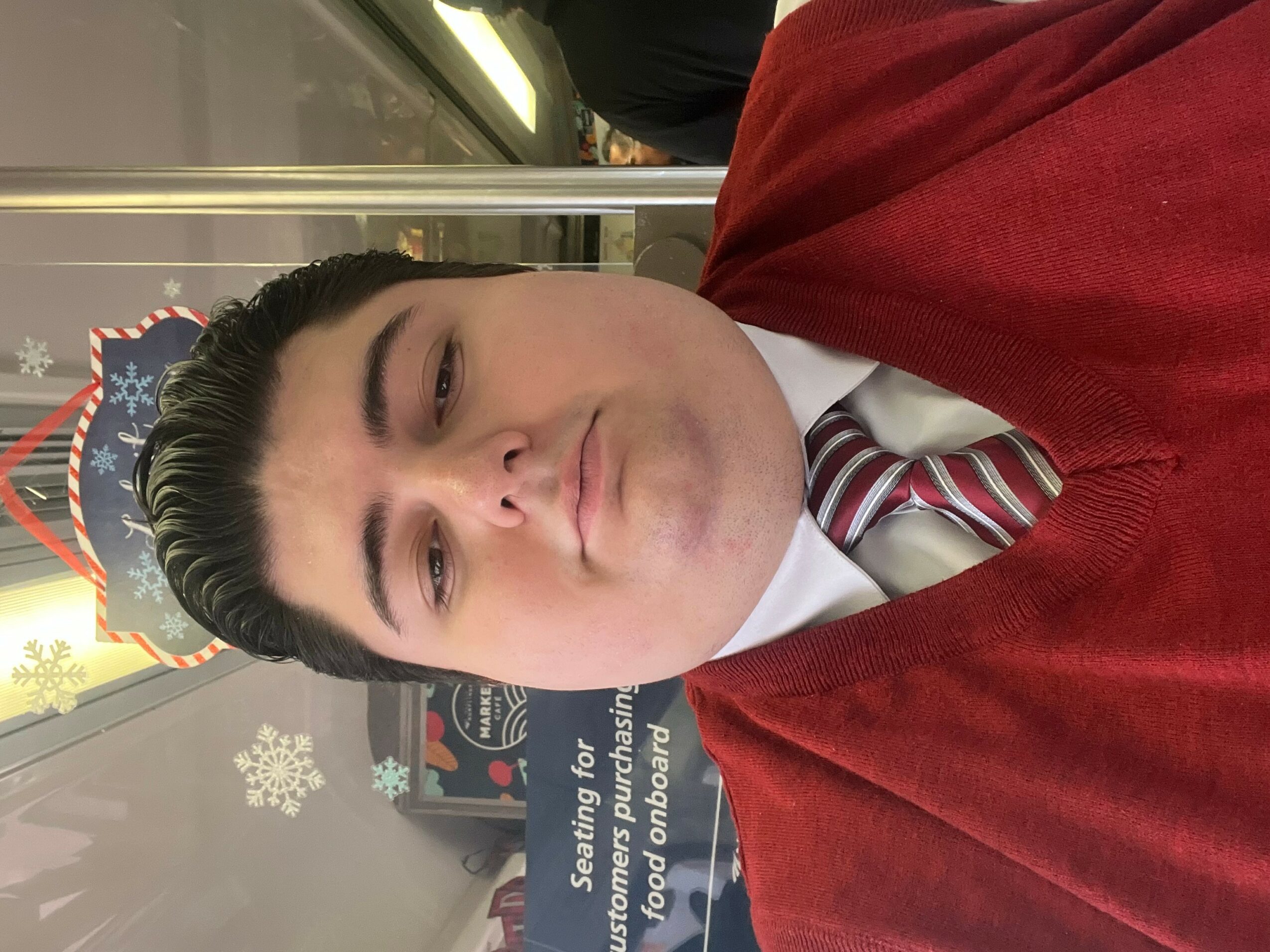 Young man in a red sweater and striped tie, looking at the camera, with a festive background behind glass.