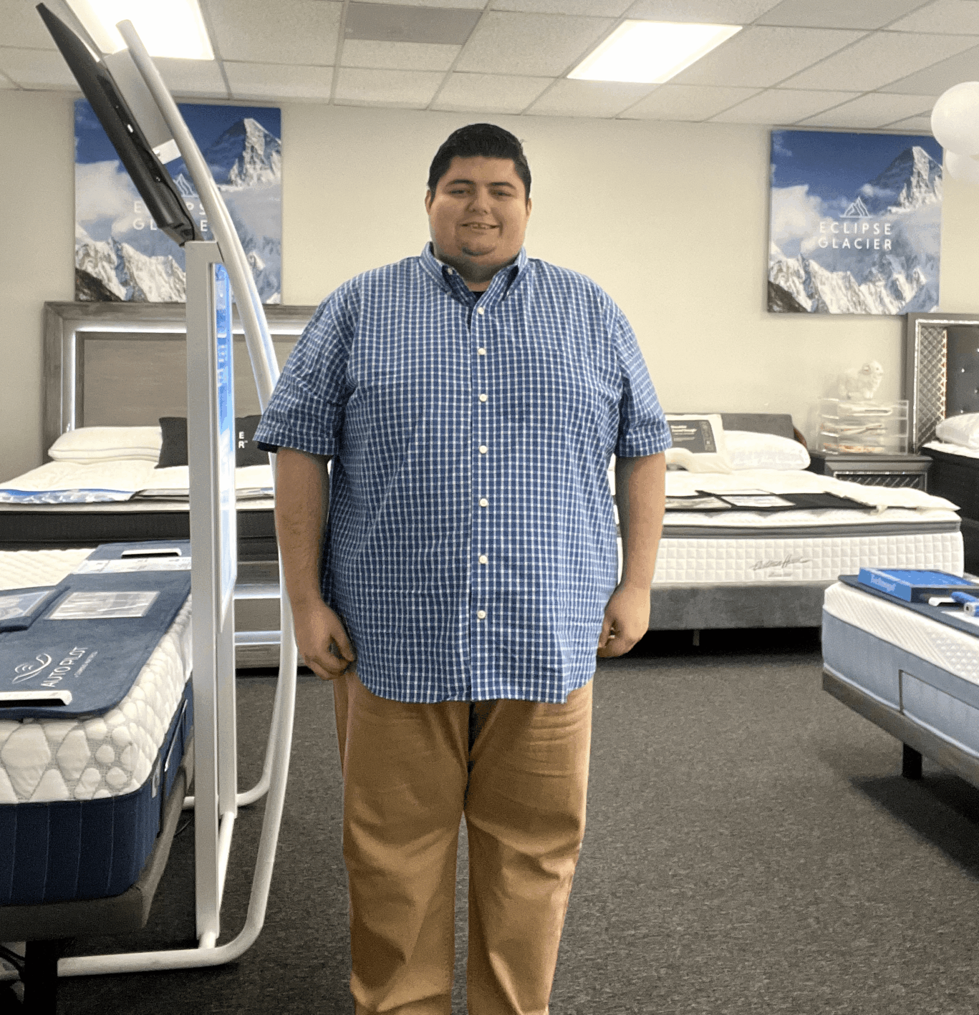A man stands in a mattress store, smiling, wearing a blue checkered shirt and khaki pants, with beds and posters in the background.