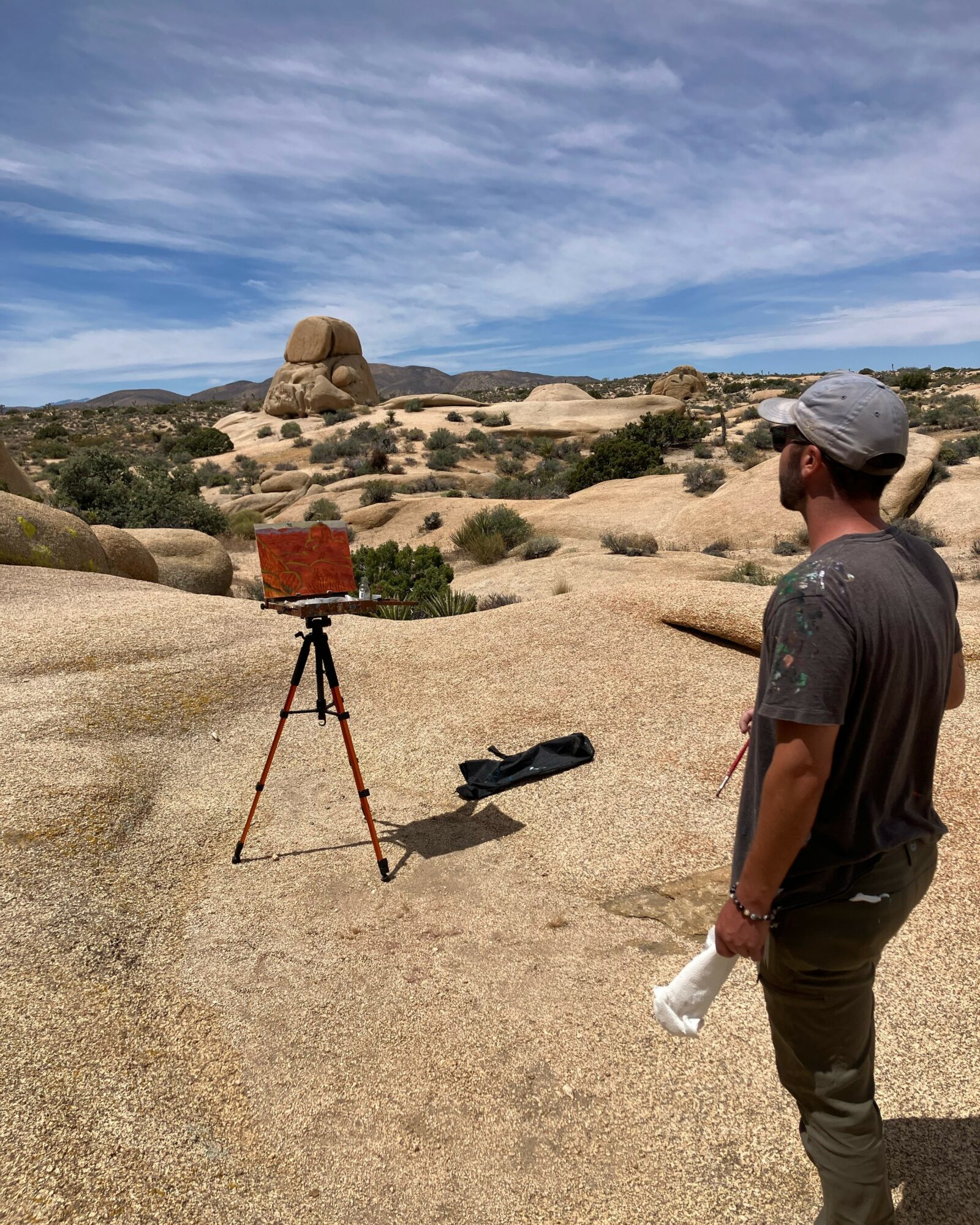 Person standing outdoors in a rocky desert landscape with a camera on a tripod, holding a water bottle, under a partly cloudy sky.