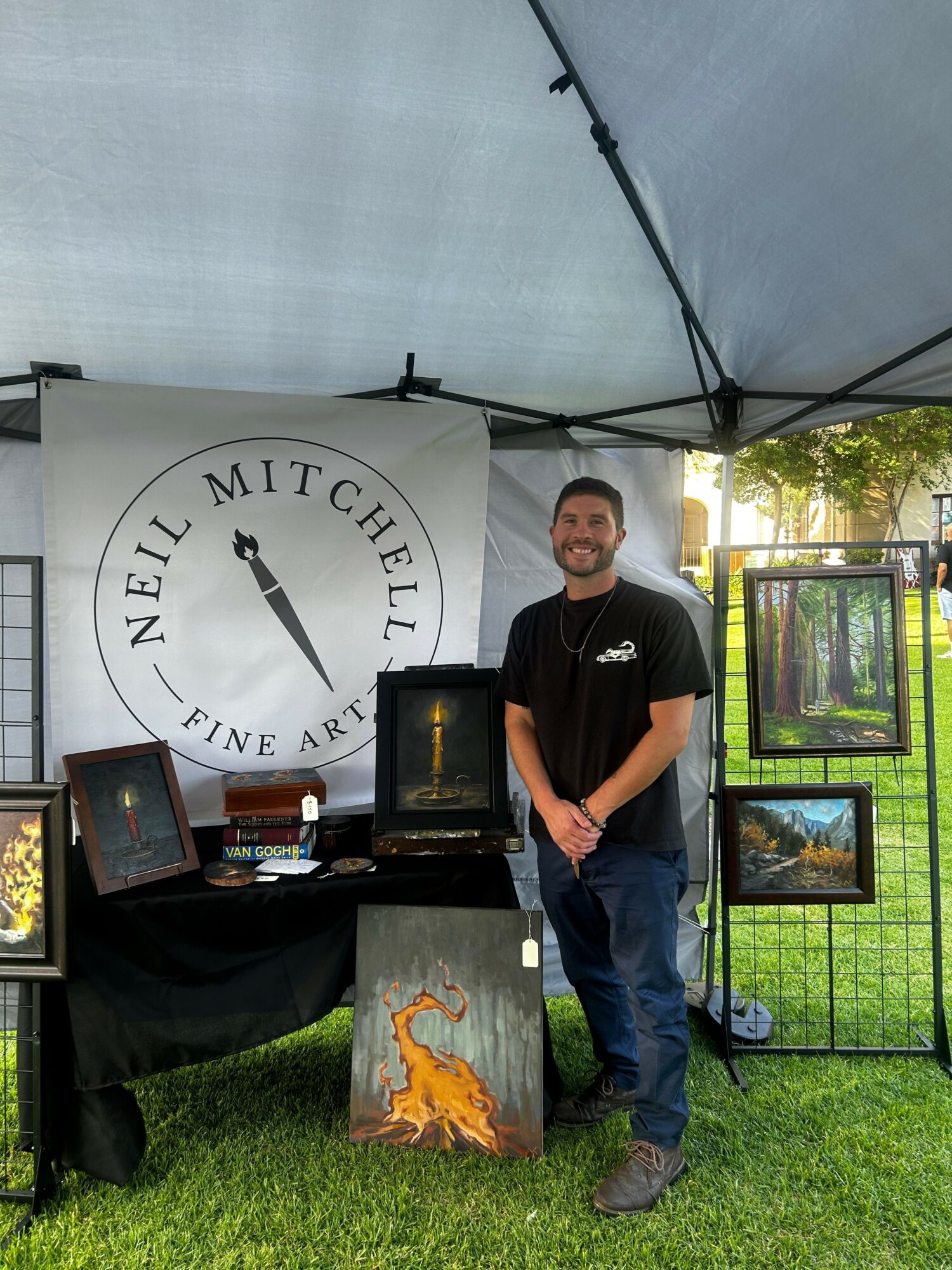 Man standing outdoors next to display with a large circular sign reading 'Mitchell Neill' and a flame sculpture, with framed artwork behind him.