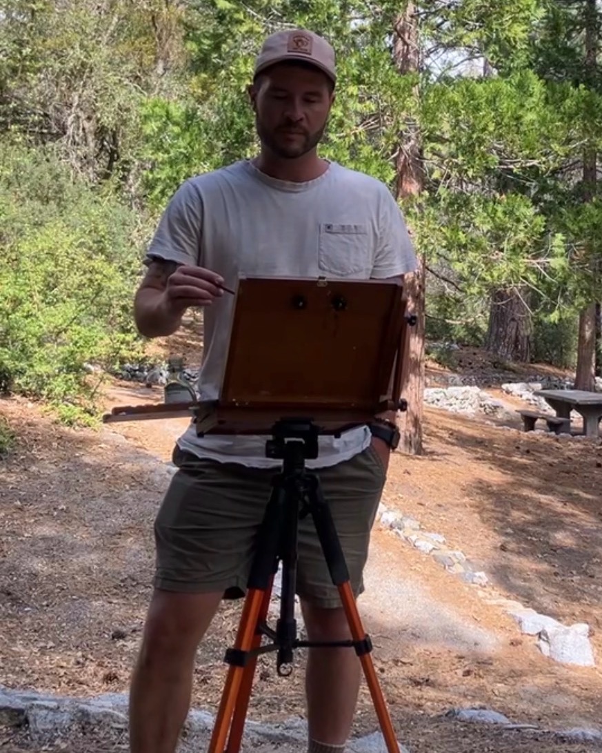 Man outdoors painting on a portable easel, surrounded by trees and nature, wearing a cap and casual clothing.