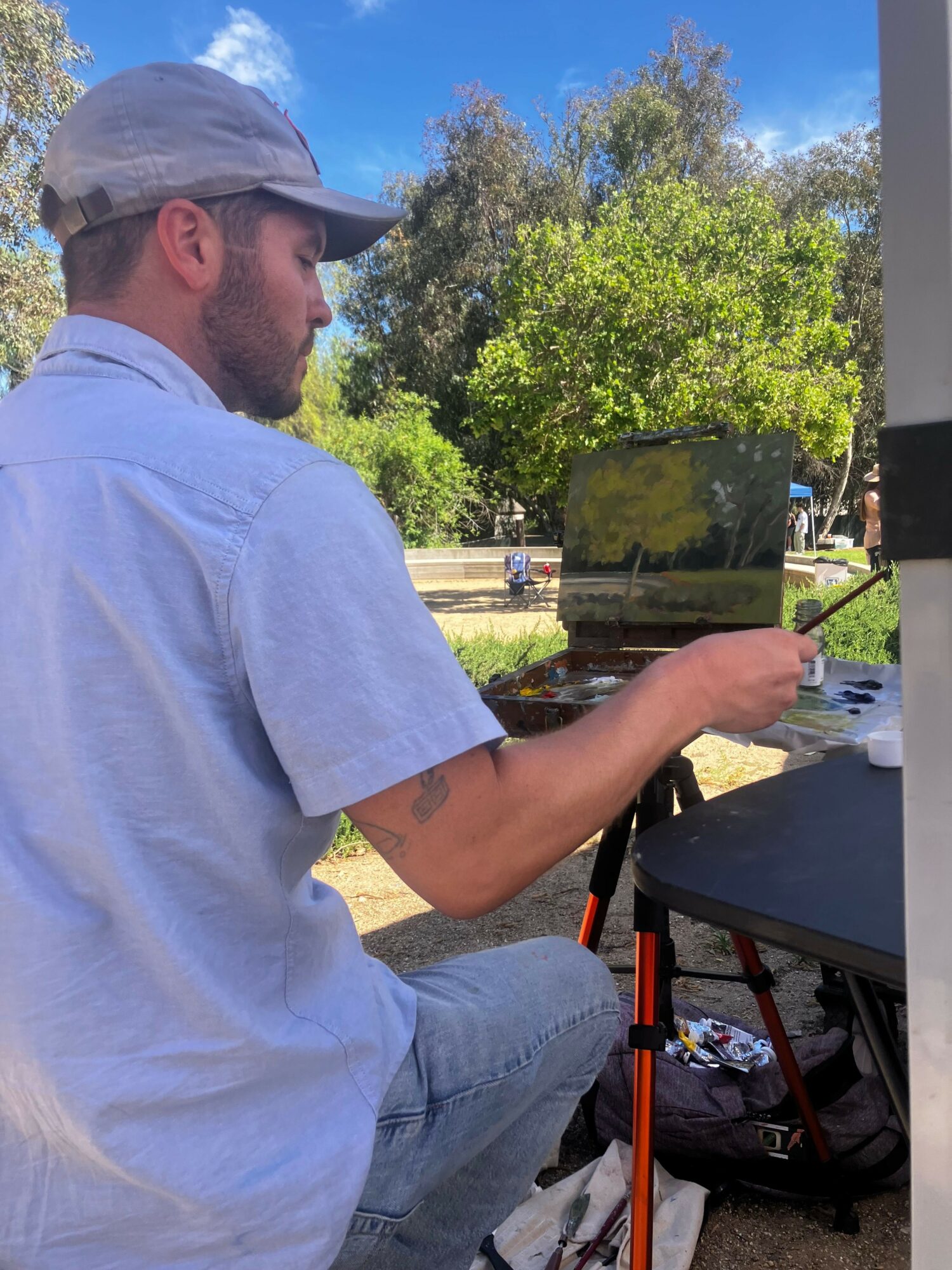Man sitting outdoors, wearing a cap and light-colored shirt, holding a drink with trees and blue sky in background.