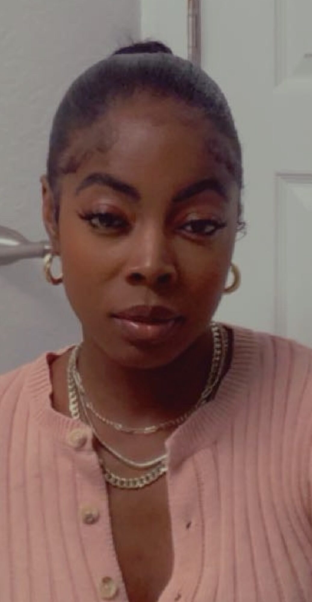 Young woman with dark hair in a high bun, wearing gold hoop earrings, layered necklaces, and a pink top, looking at the camera.