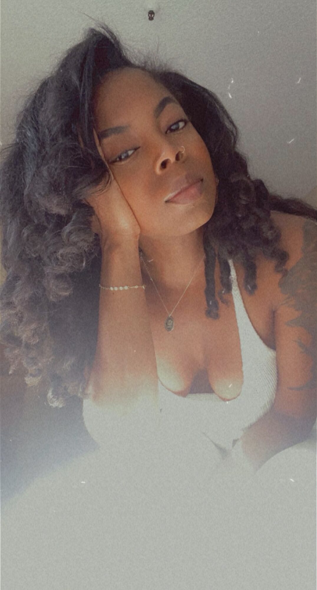 Young woman with curly hair resting her head on her hand, wearing a white top and jewelry.