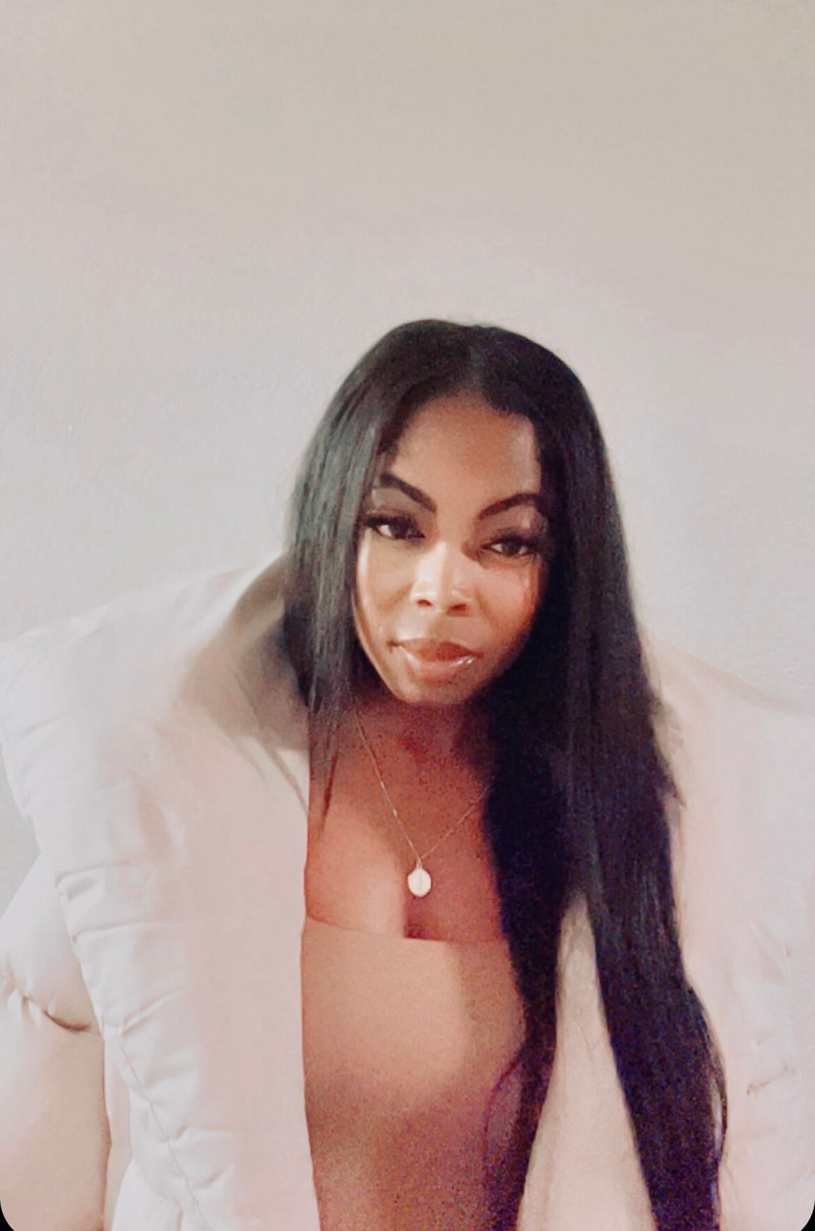 Woman with long black hair and makeup, wearing a beige top and necklace, looking at the camera.